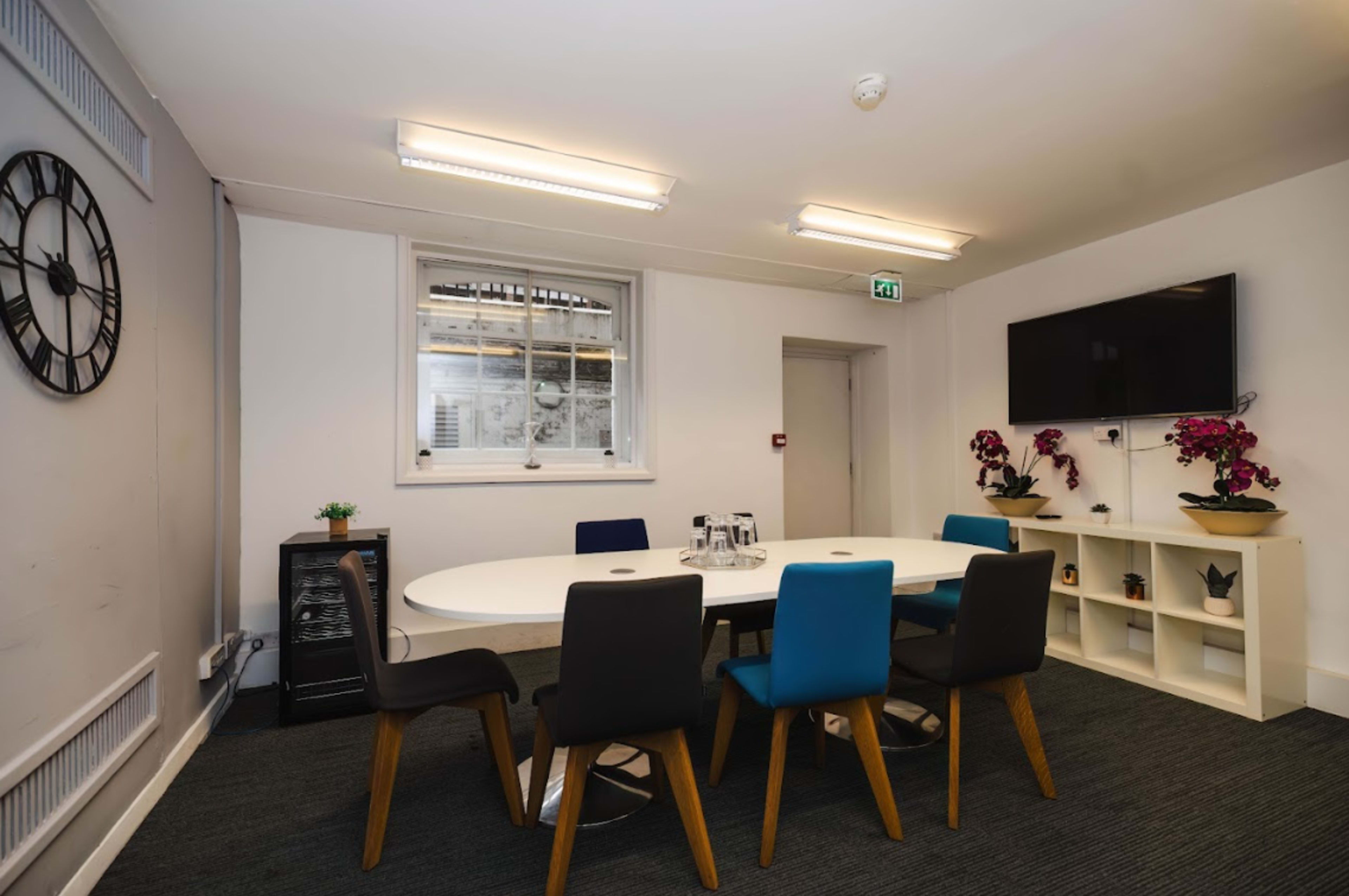 A meeting room features a round table surrounded by chairs, a wall-mounted television, and a window with plants on the sill.