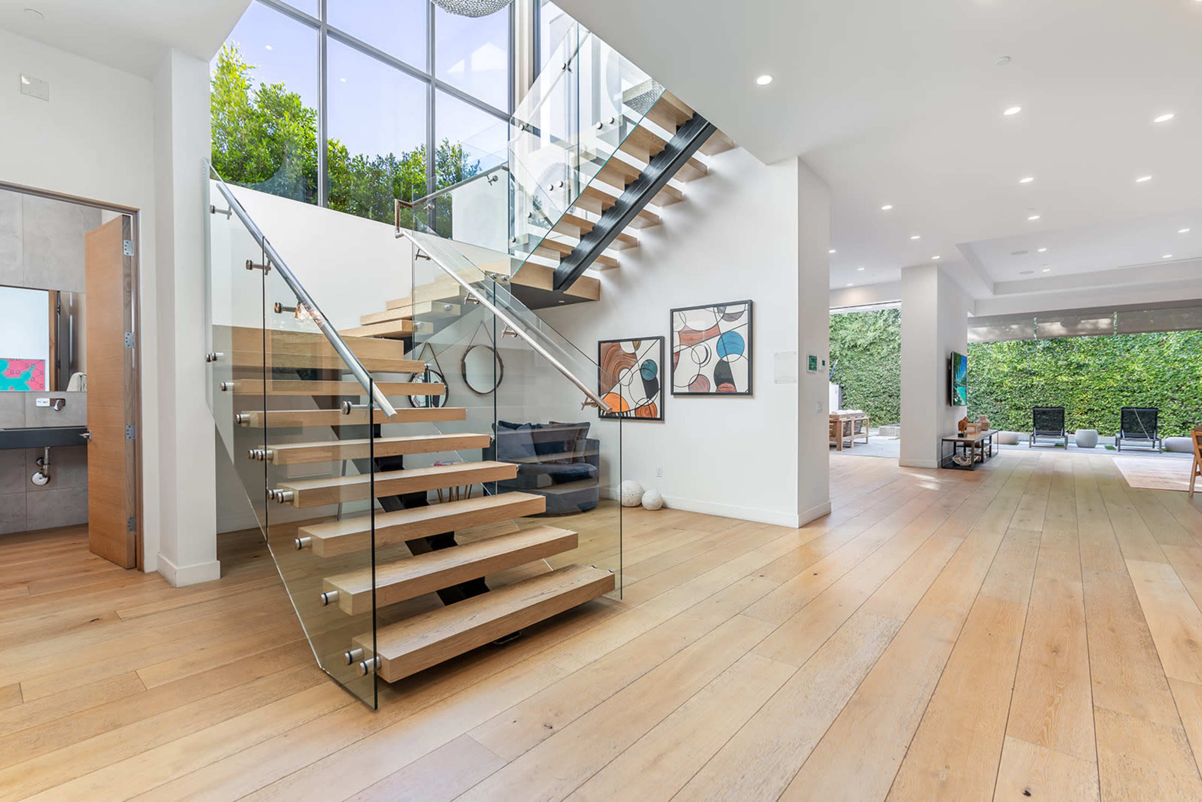 A modern interior featuring a staircase with wooden steps and a glass railing, leading to an upper level, with artwork displayed on the walls and large windows illuminating the space.