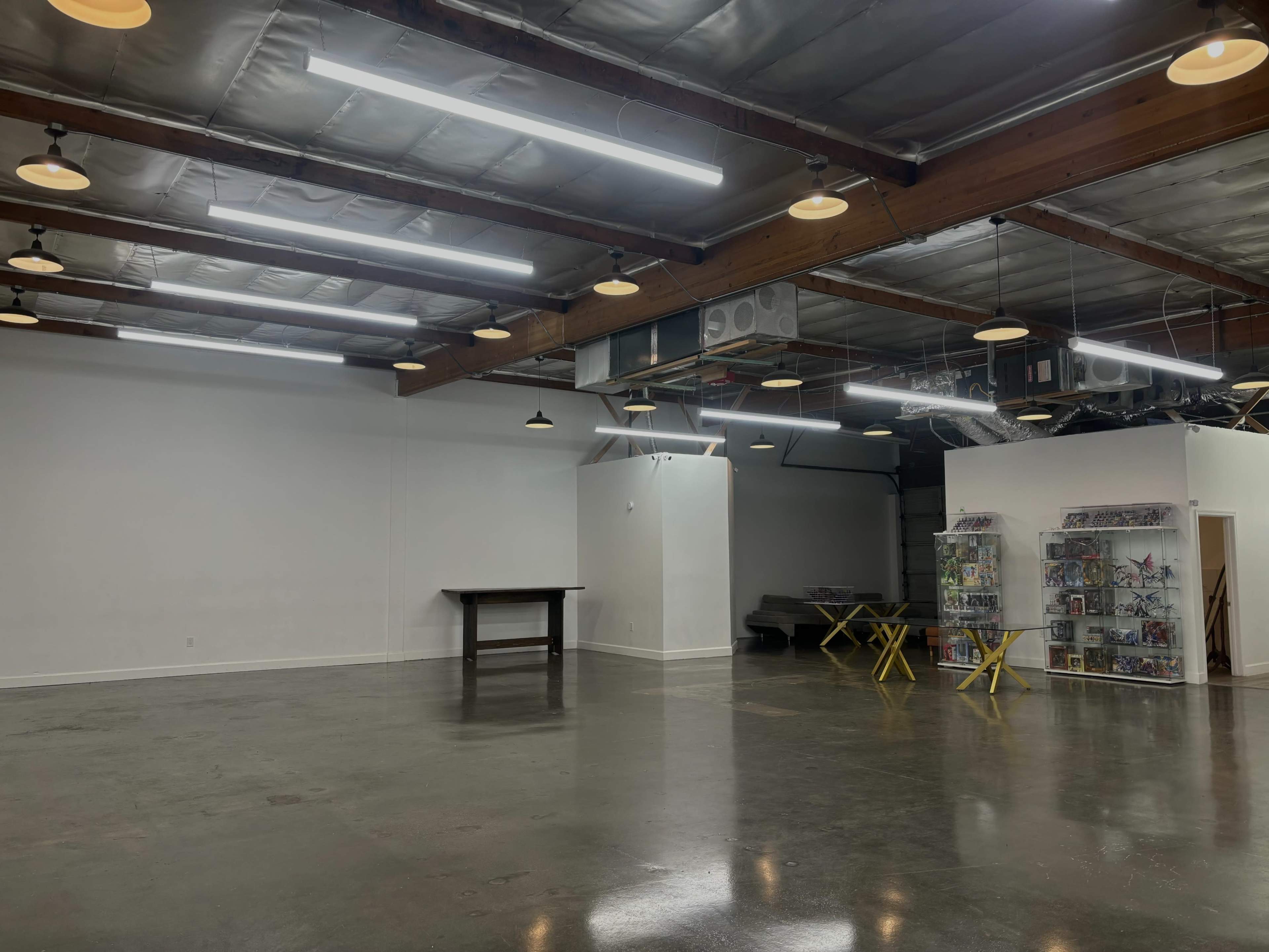 The image shows a spacious, empty room with polished concrete floors, exposed beams, overhead lighting, and a central table surrounded by a few folding chairs.