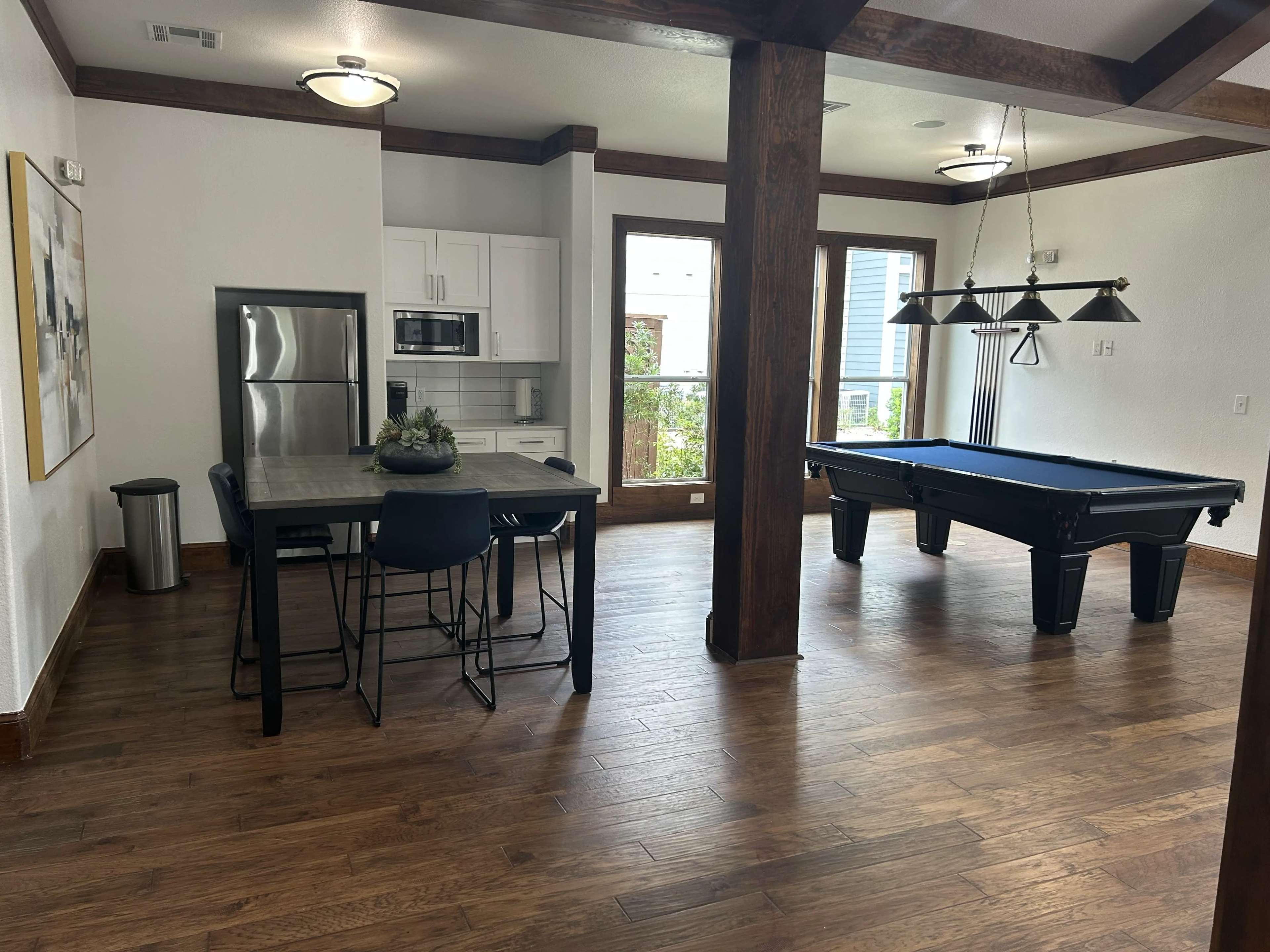 The image shows a dining area with a kitchen space featuring a stainless steel refrigerator and a black billiard table in a room with wooden flooring and modern decor.