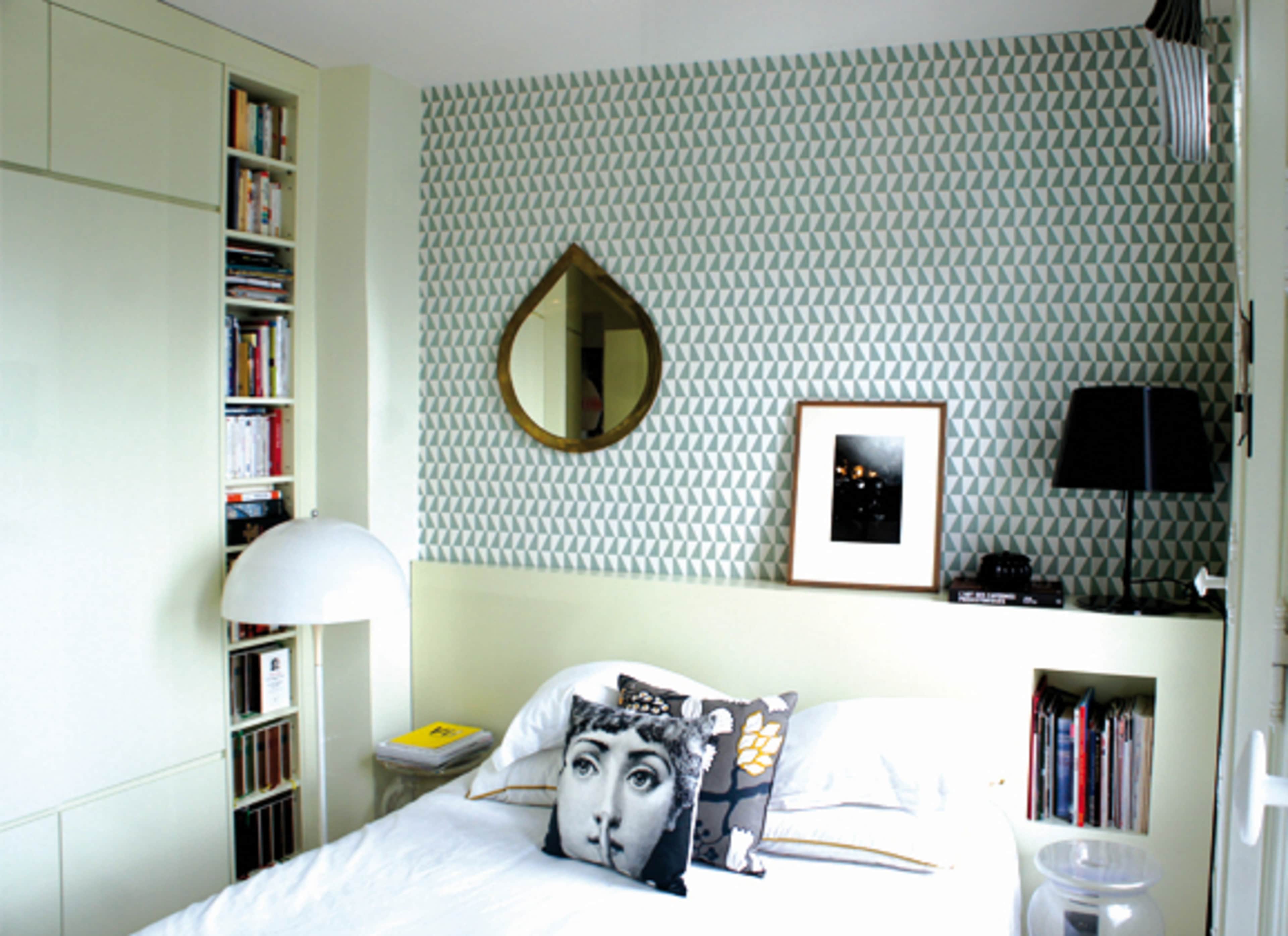 The image shows a neatly arranged bedroom featuring a bed with decorative pillows, a bookshelf, a round mirror, and a bedside lamp against a patterned wall.