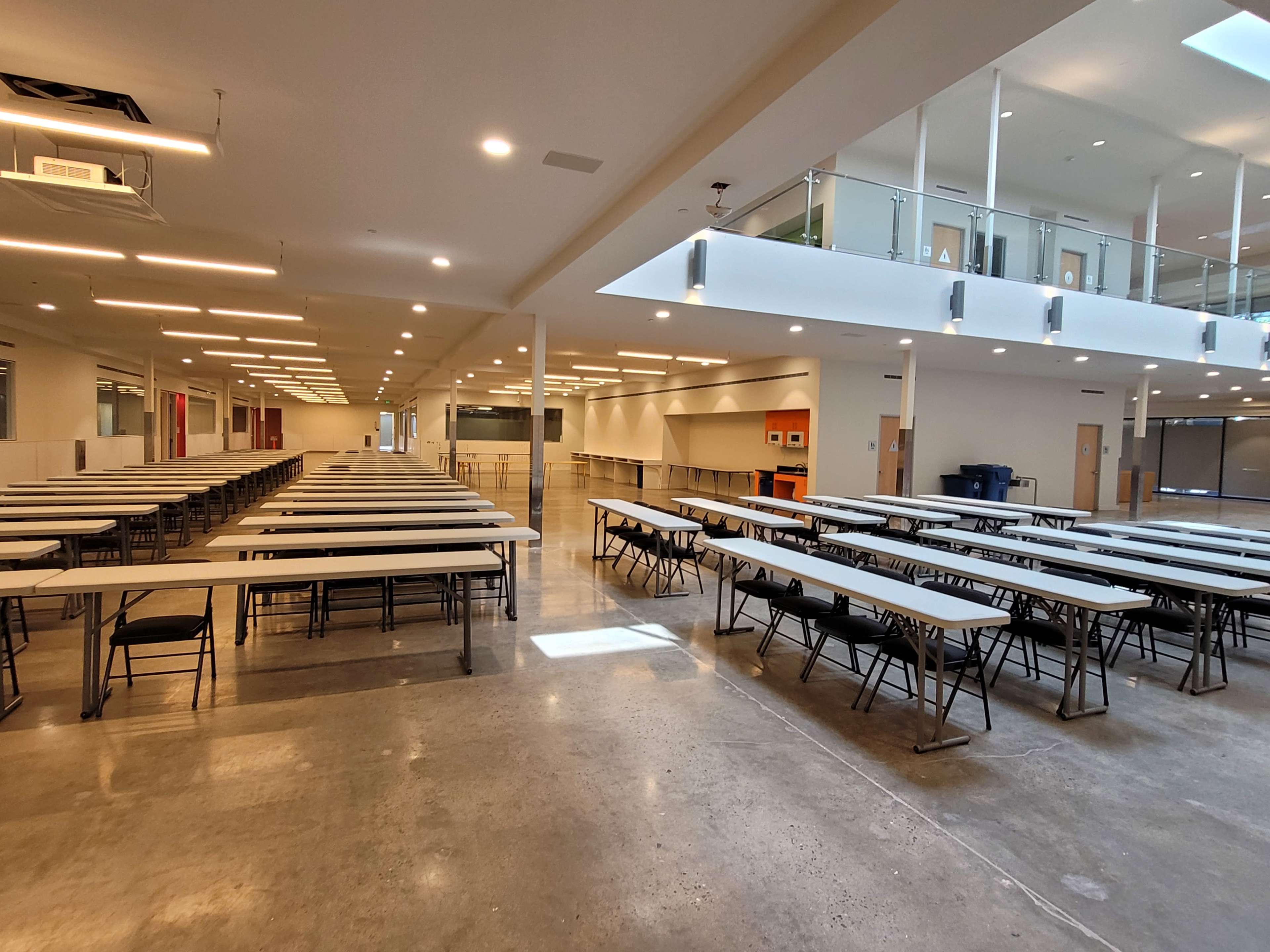The image shows a spacious indoor setting with rows of folding tables and chairs arranged in a large, brightly lit room.