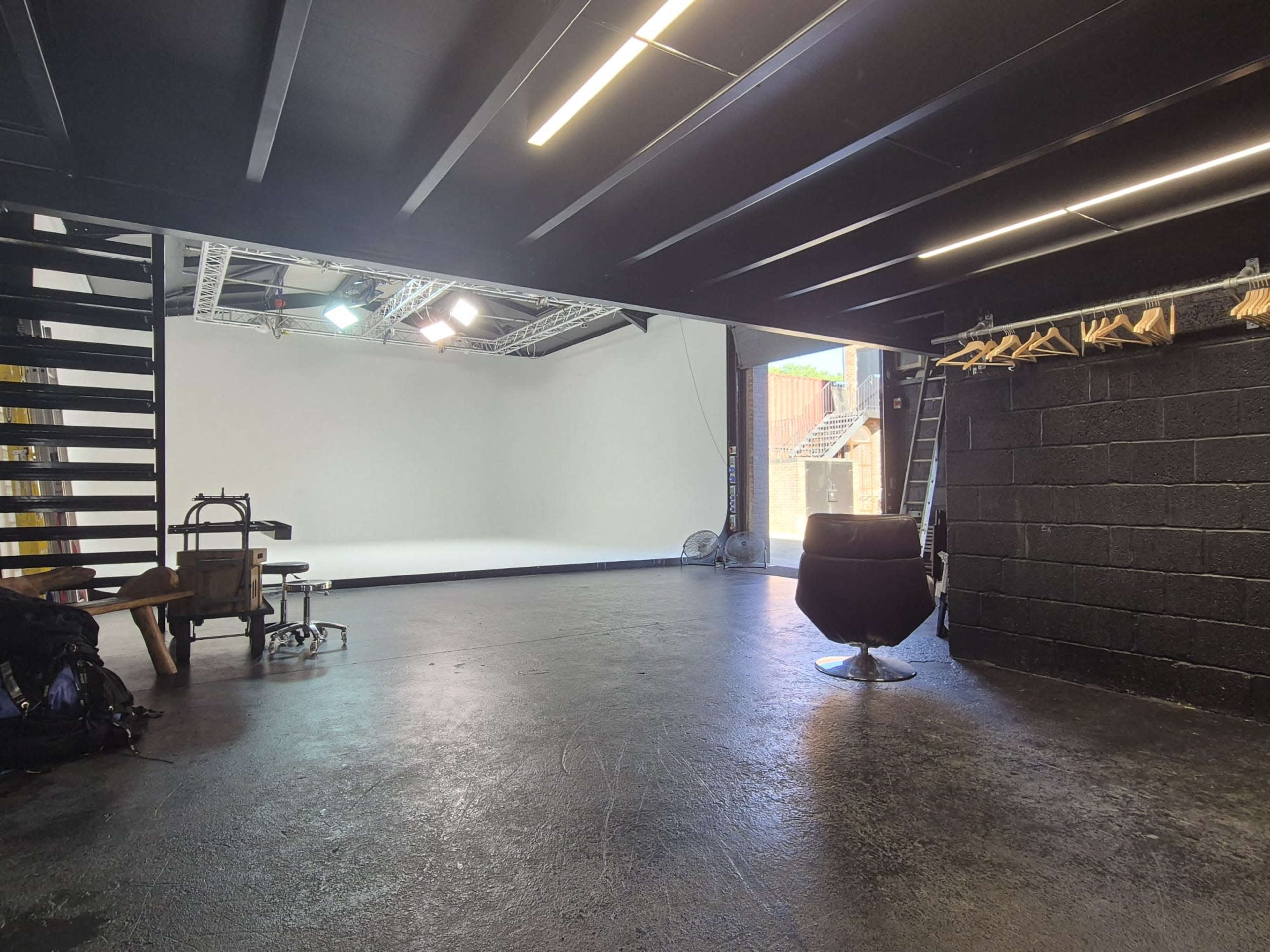 The image shows an empty studio space with a dark floor, a large white backdrop, and a chair positioned in front of it, along with an adjacent staircase and some equipment on the side.