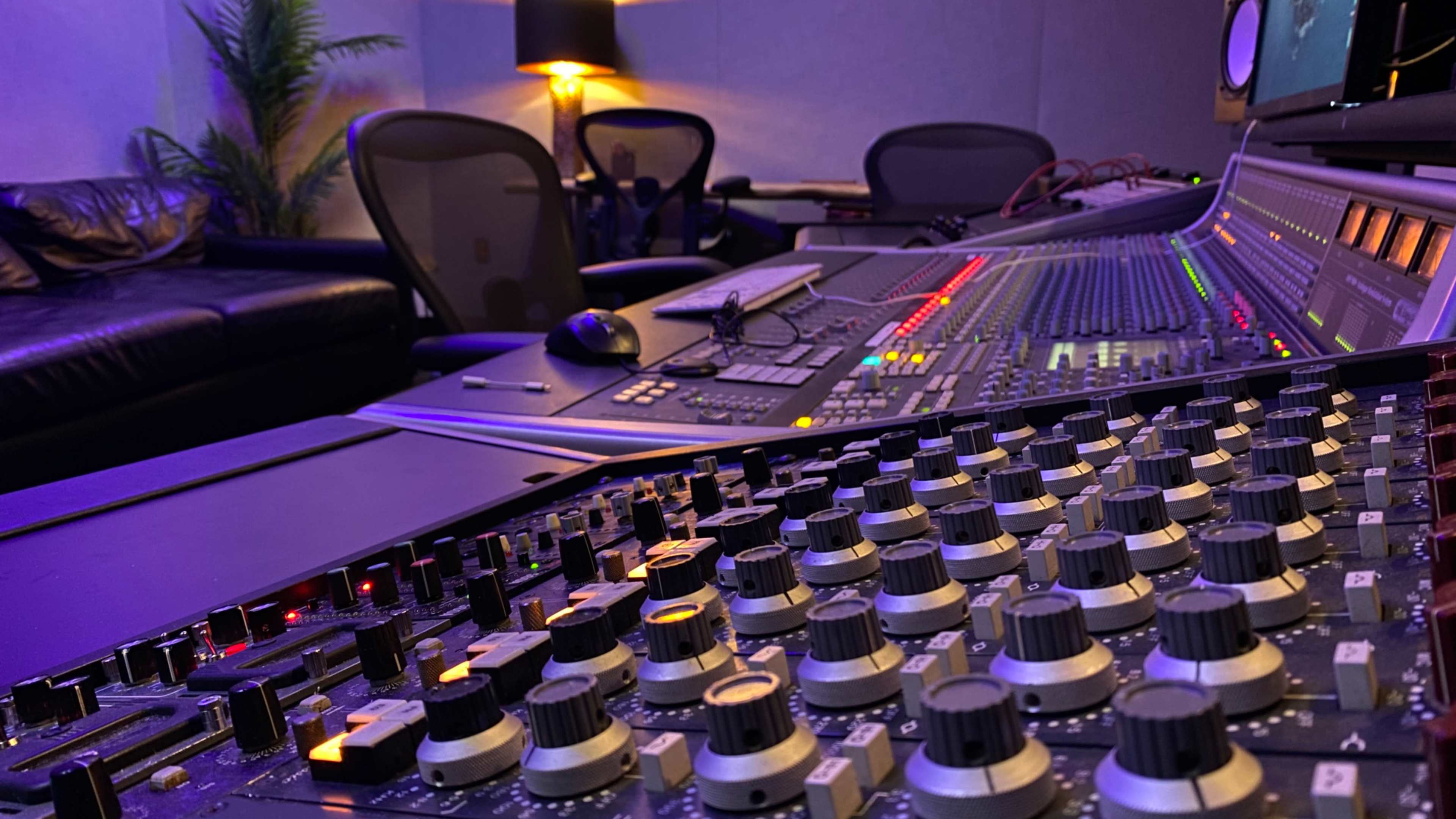 The image shows a close-up of a mixing console with various knobs and sliders in a dimly lit recording studio.