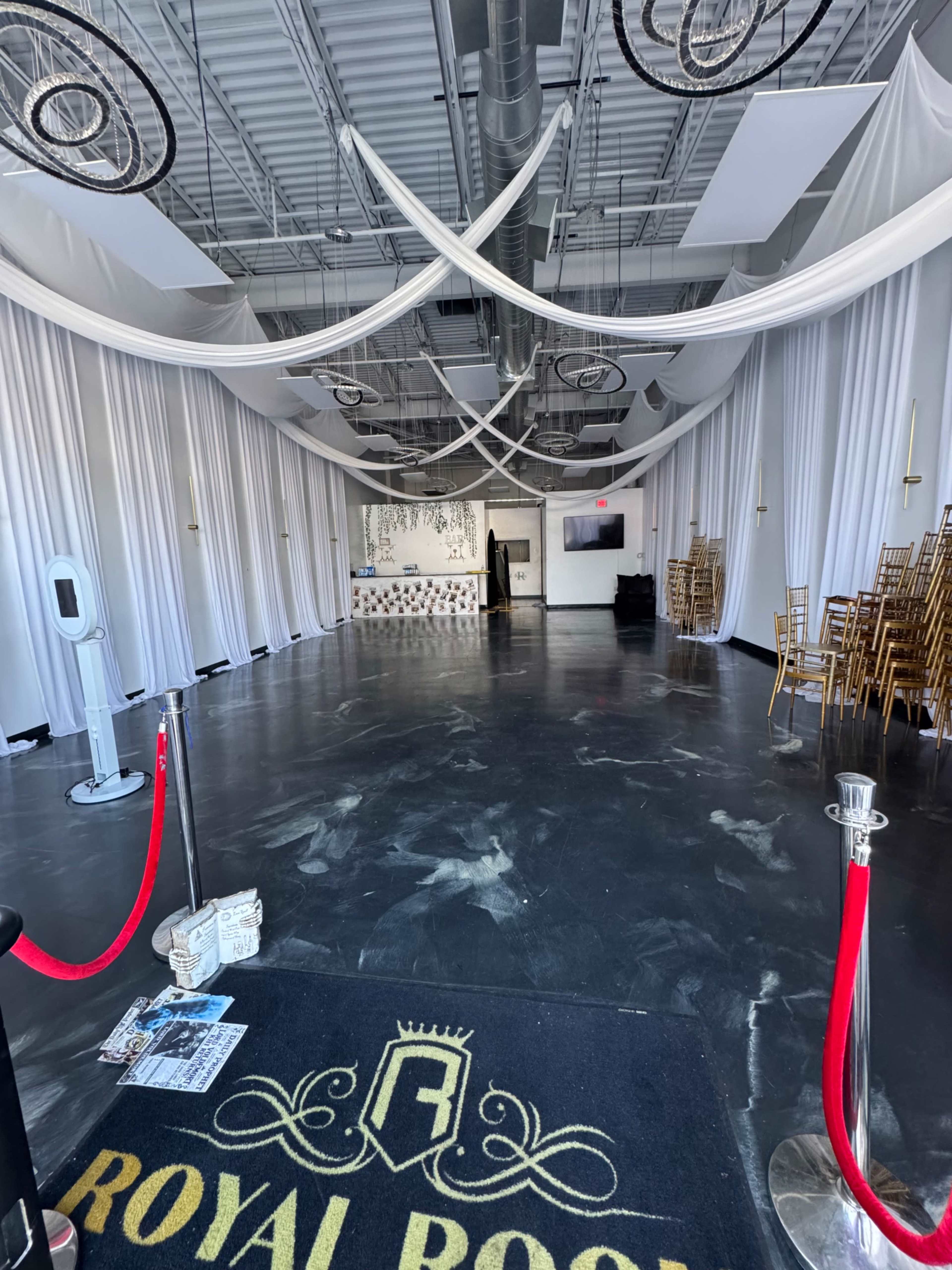 The image shows an empty event space with white curtains, a reception area, and gold chairs stacked against the wall.