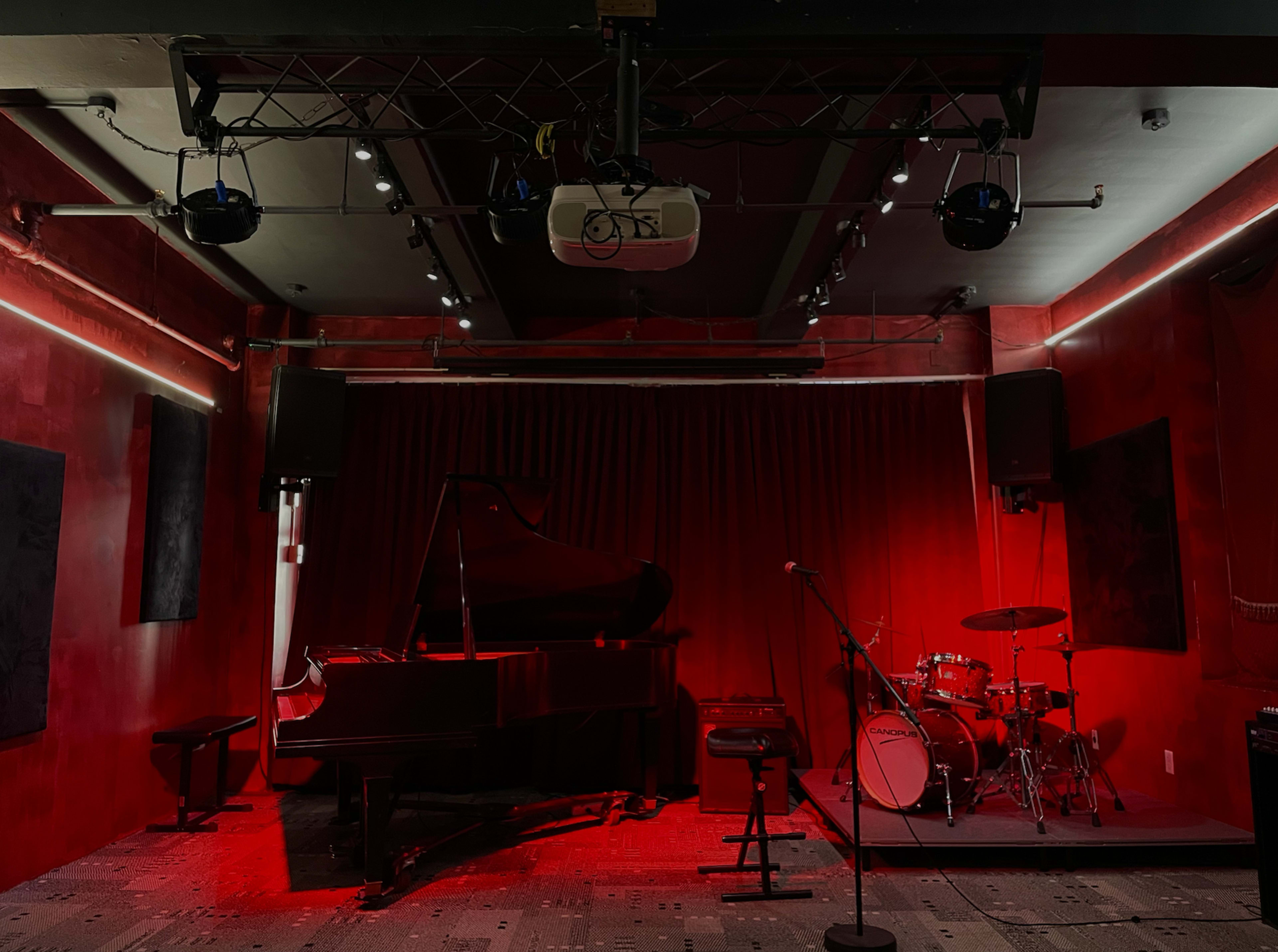 A dimly lit music venue features a grand piano, a drum set, and red walls with sound equipment mounted above.