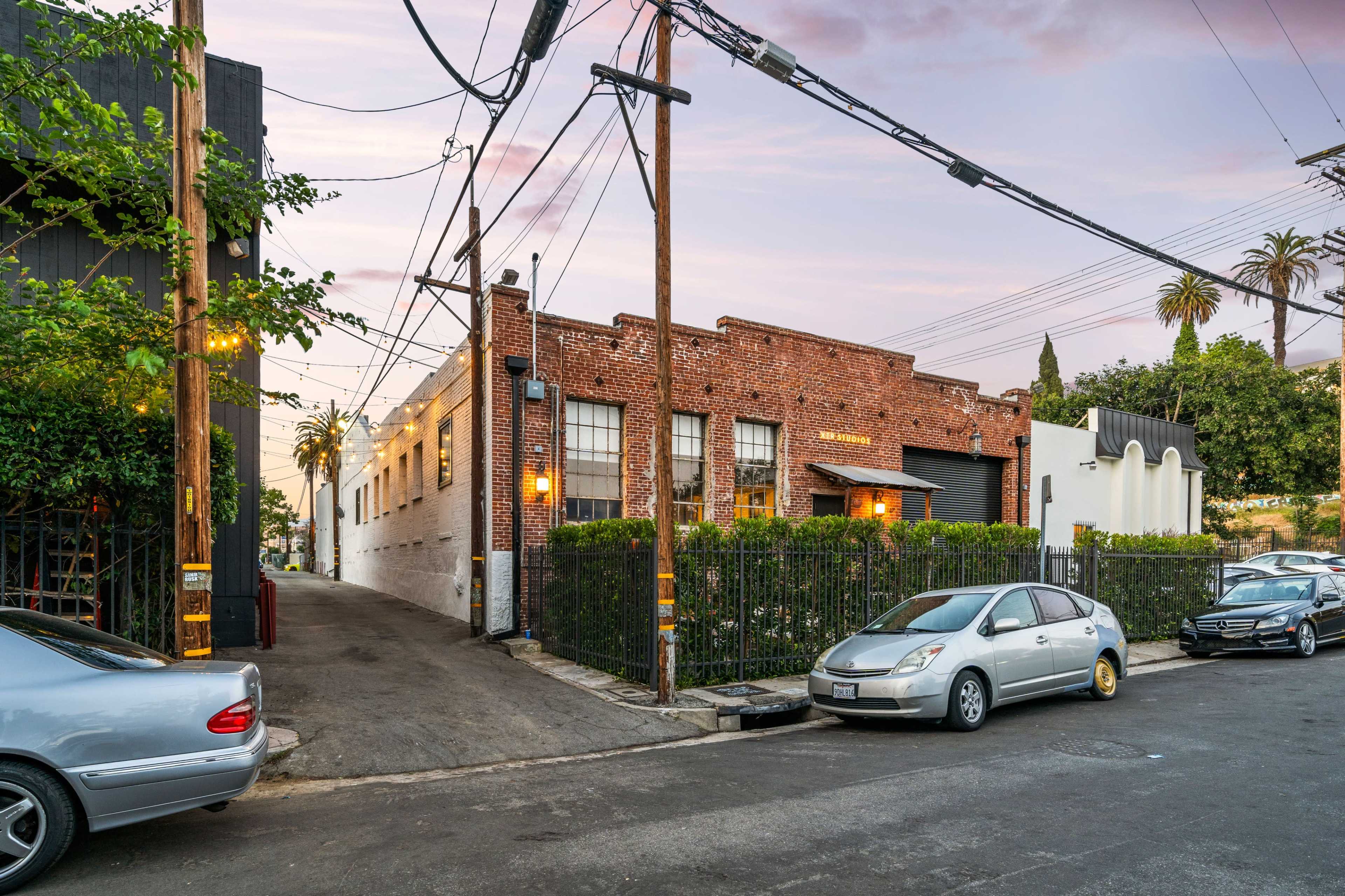 A brick building with large windows is situated at the end of a narrow alley flanked by parked cars and power lines under a pastel sky.