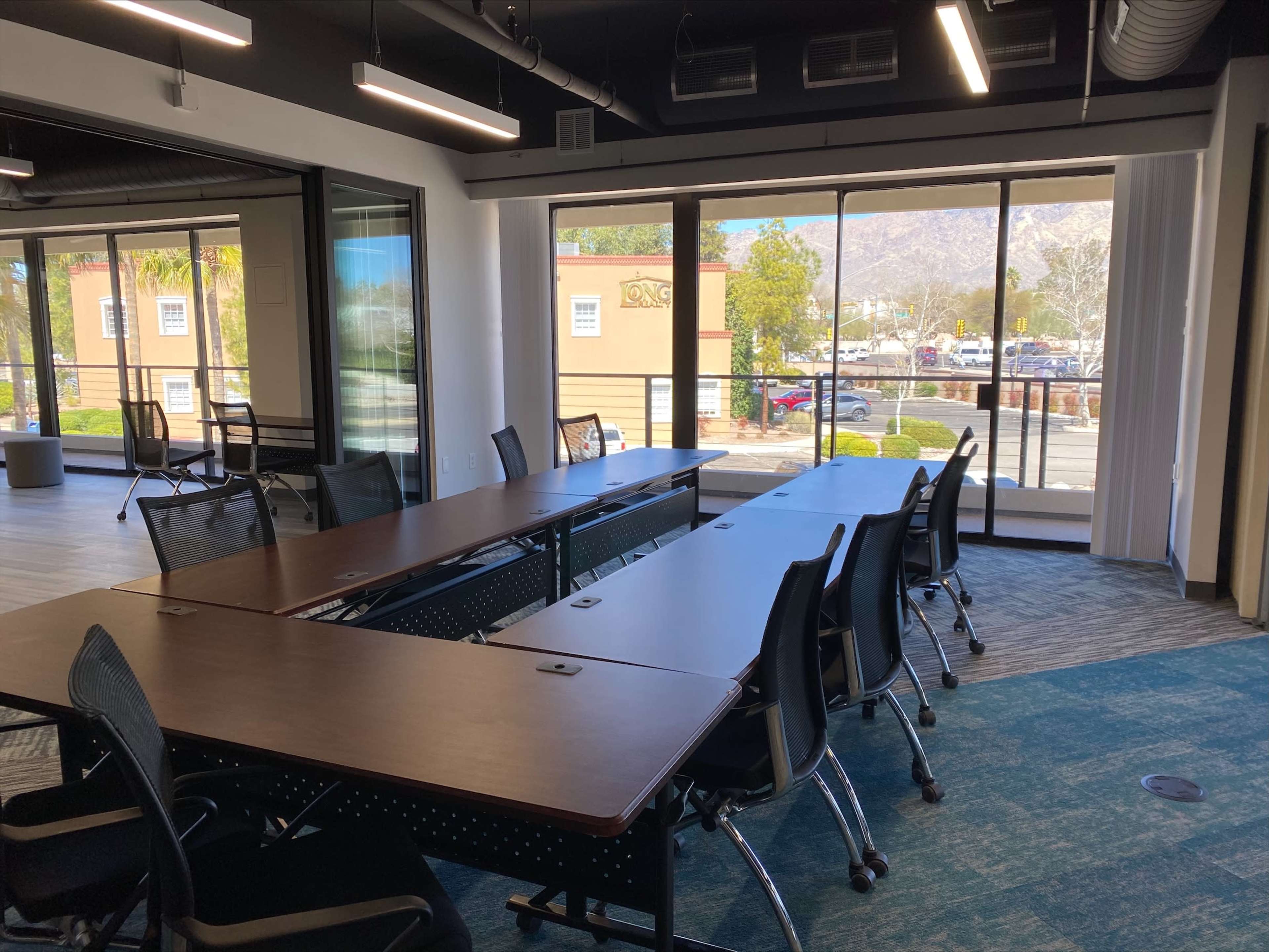 A conference room with several tables arranged in a U-shape and a large window that offers a view of a parking lot and mountains in the background.