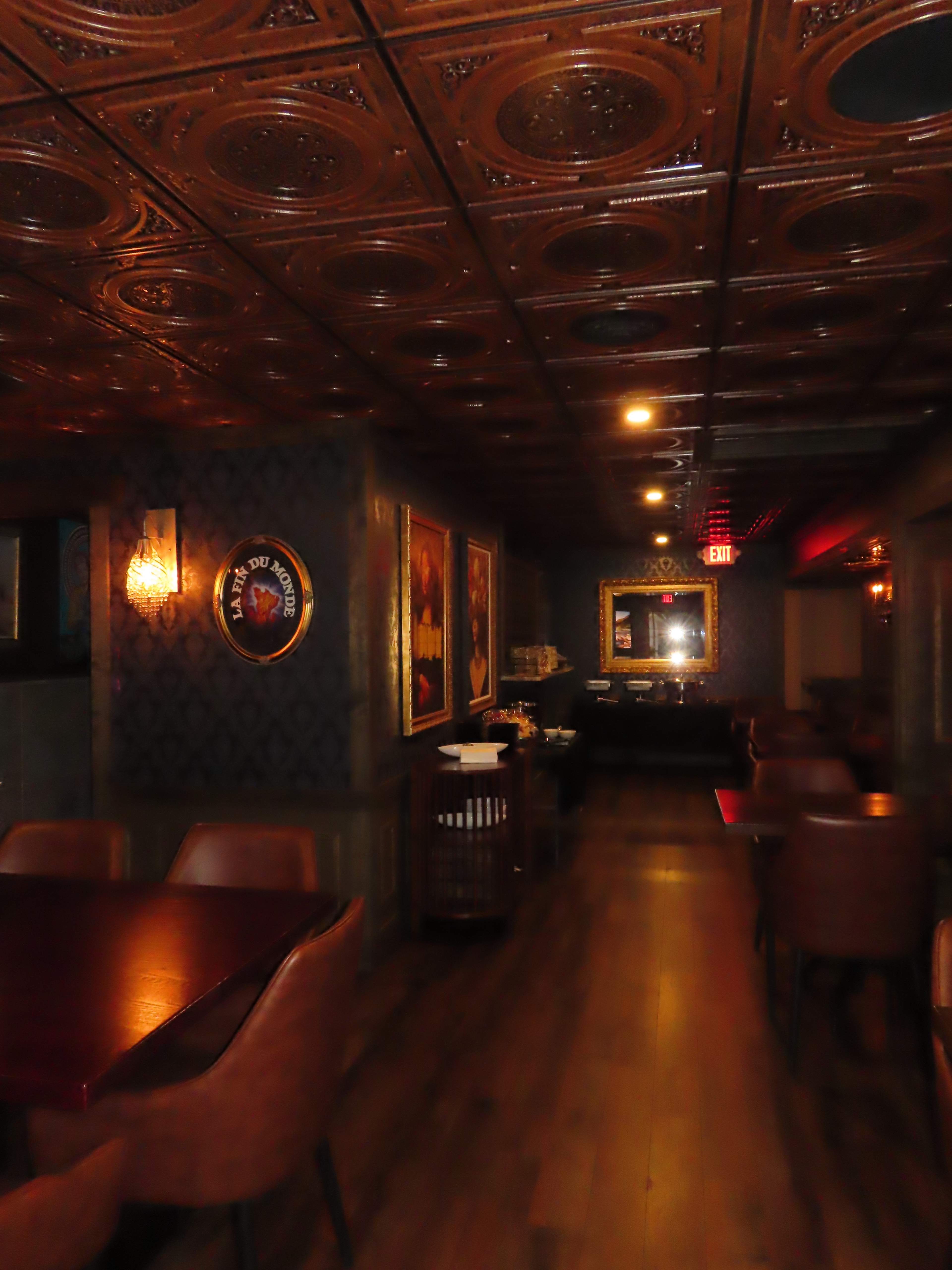 The image shows a dimly lit restaurant interior featuring wooden floors, ornate ceiling tiles, and tables arranged along the sides.