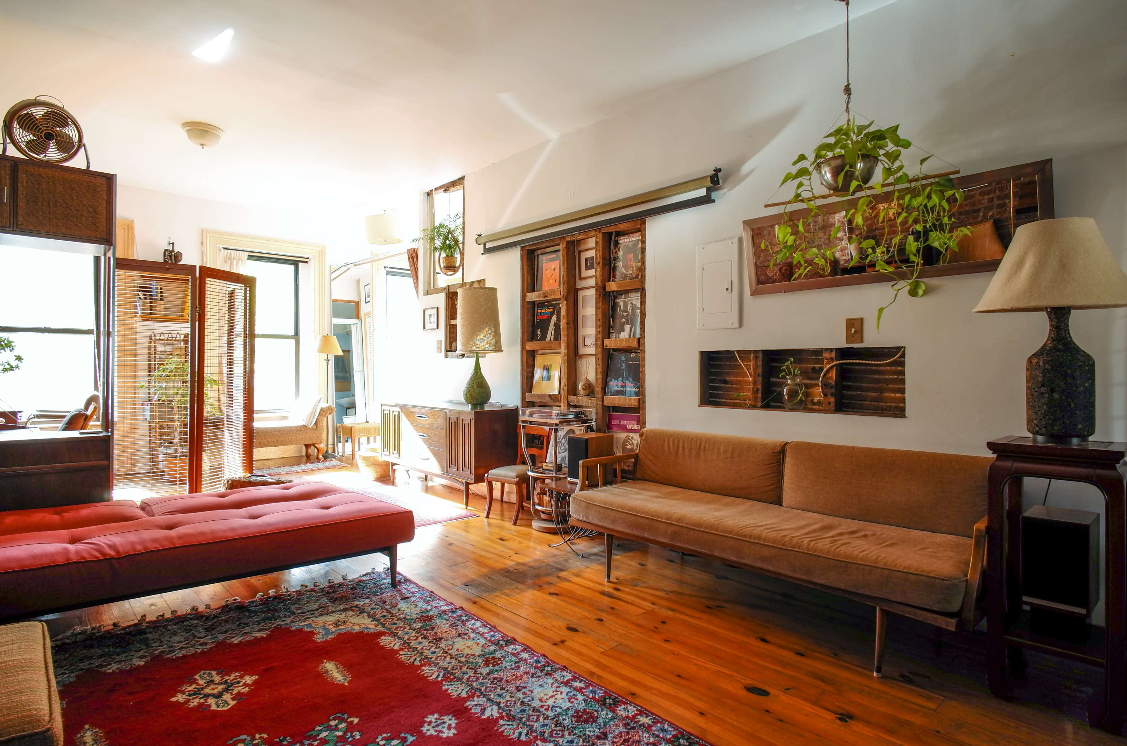 The image shows a brightly lit living room featuring vintage furniture, a red daybed, a brown sofa, and various plants and decorative items against wooden flooring.