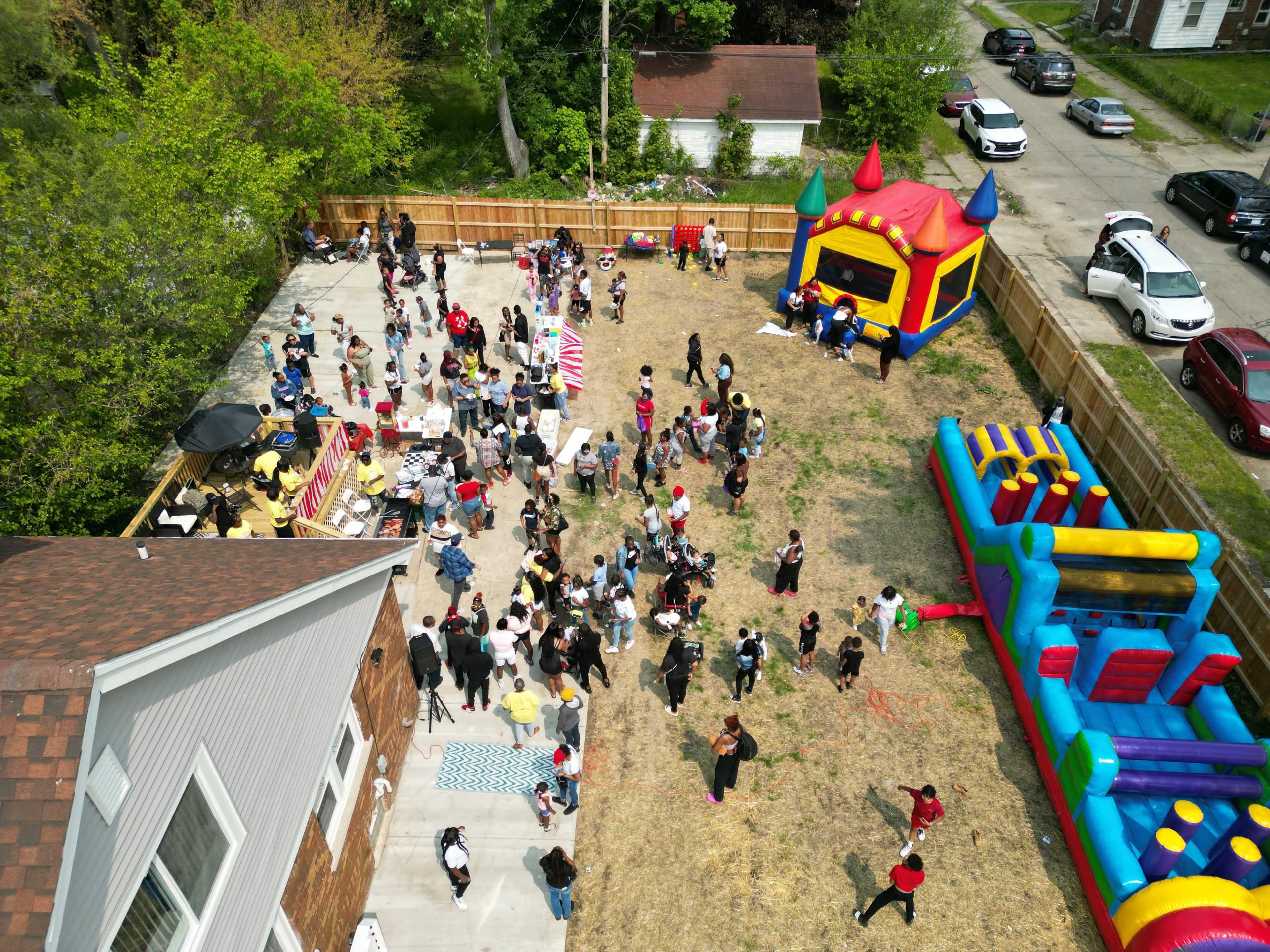 A large group of people gathers in a backyard filled with a bounce house, food stalls, and festive activities.