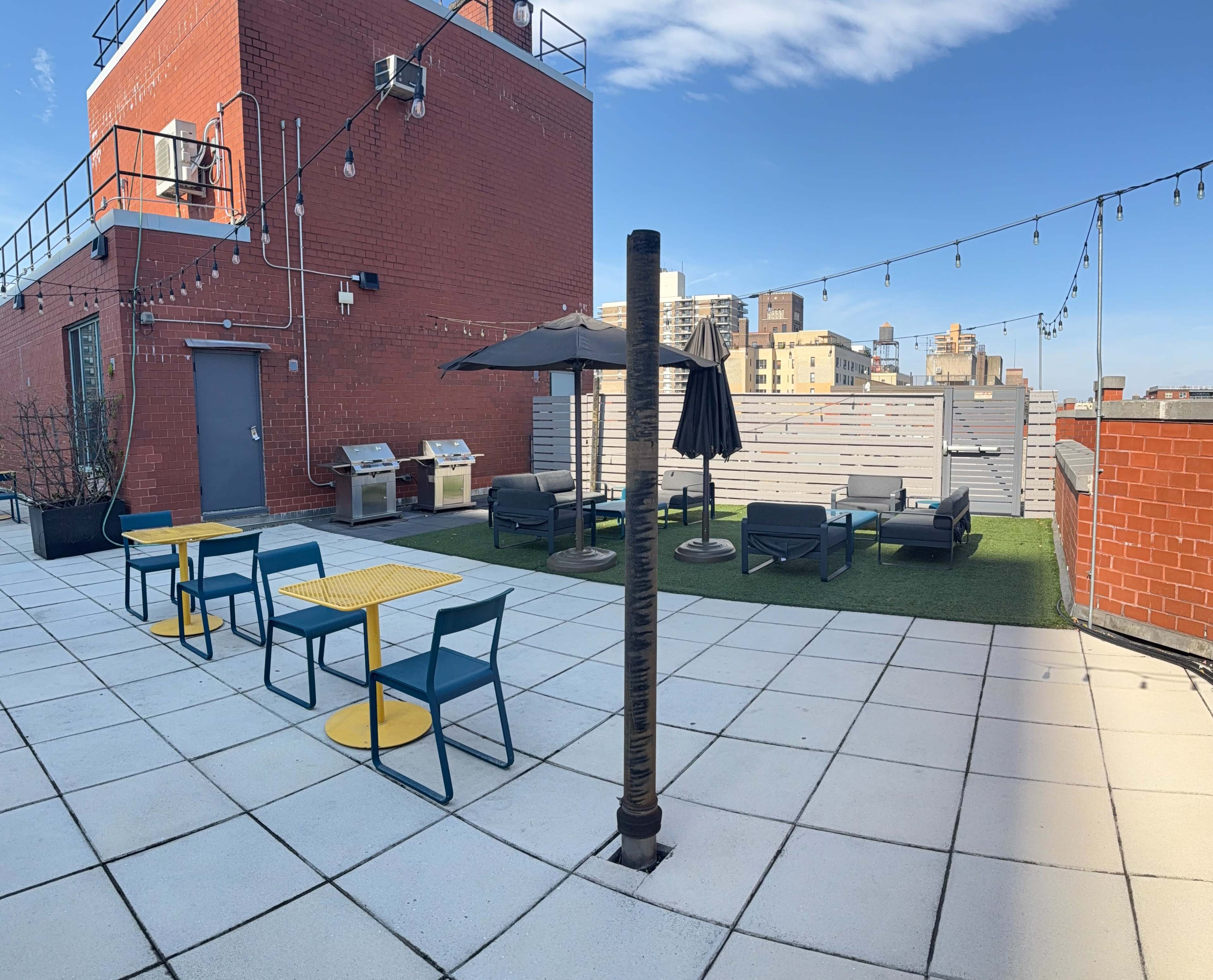 The image shows a rooftop patio with outdoor seating, grills, and a parasol, surrounded by city buildings under a clear sky.
