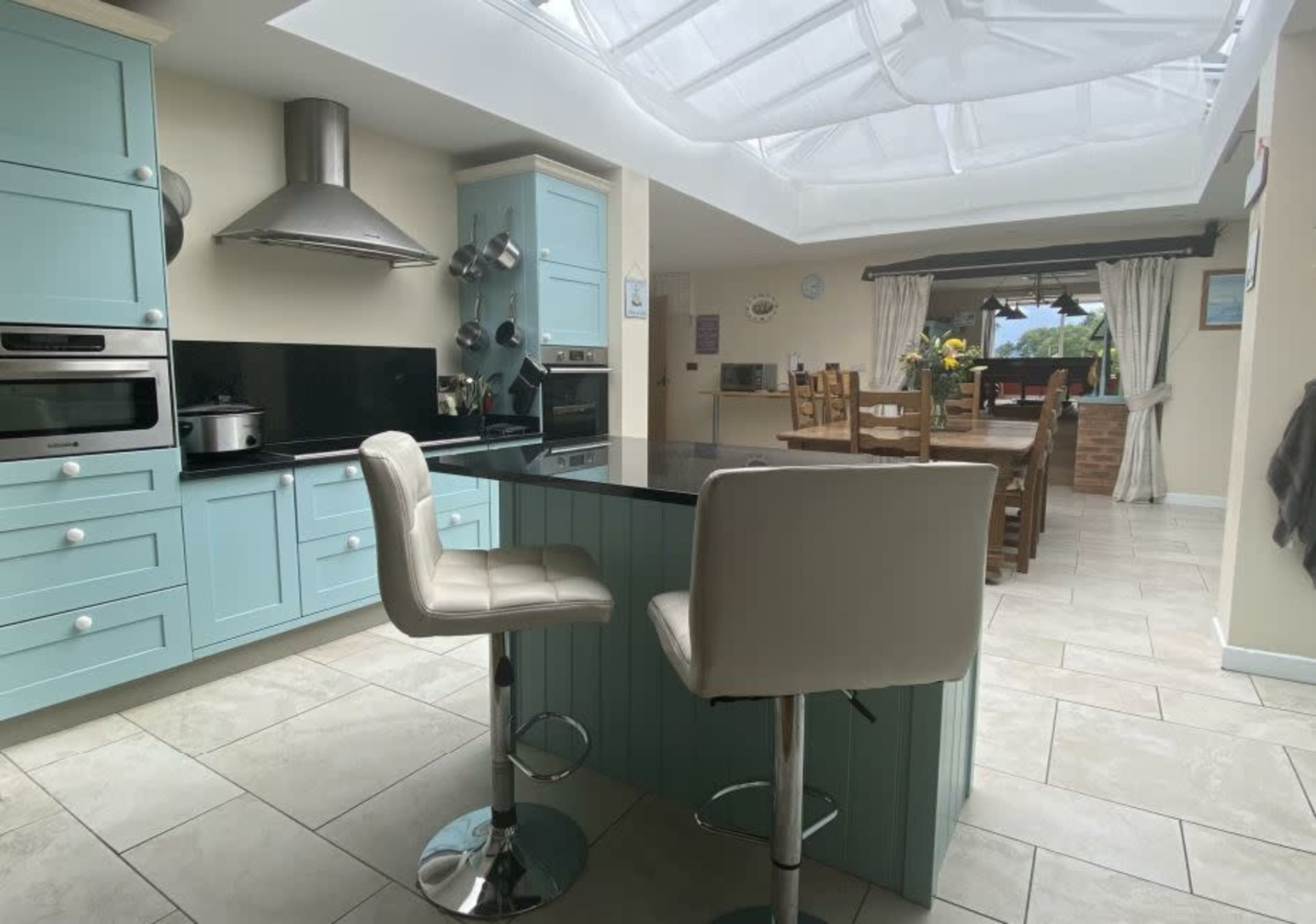 The image shows a modern kitchen featuring light blue cabinetry, a black countertop, and a seating area with two white bar stools, all illuminated by natural light from a skylight.