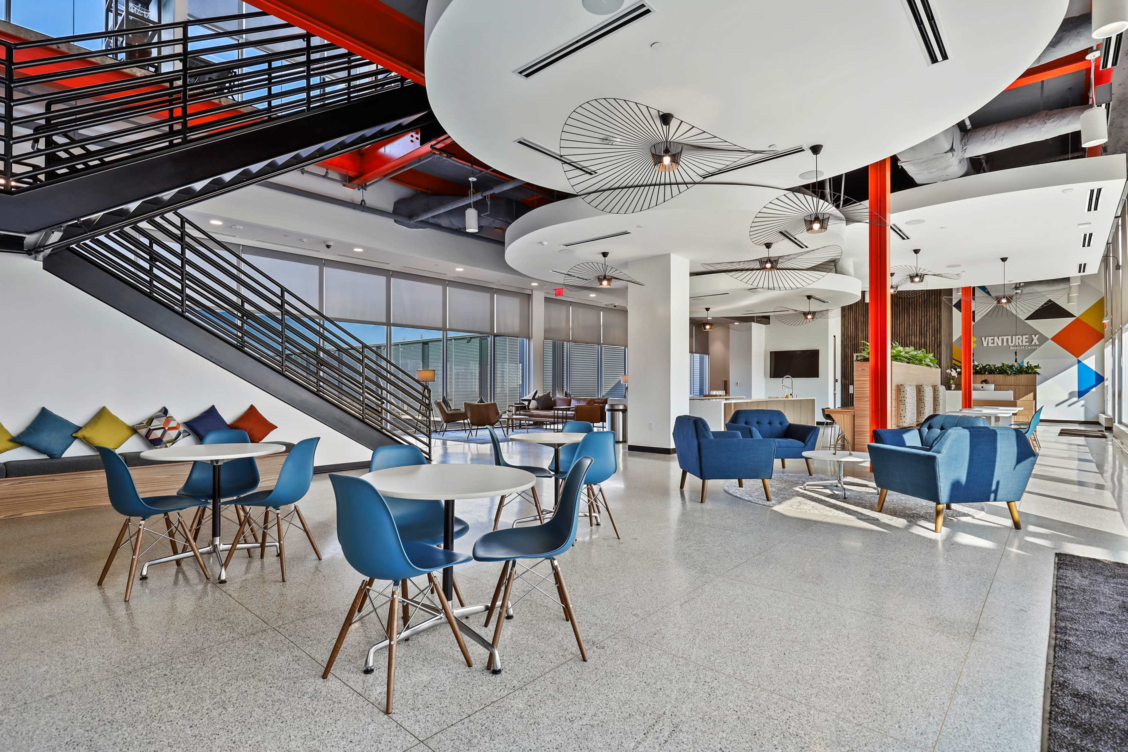 A modern office space featuring a seating area with blue chairs and tables, a staircase, and large windows allowing natural light.