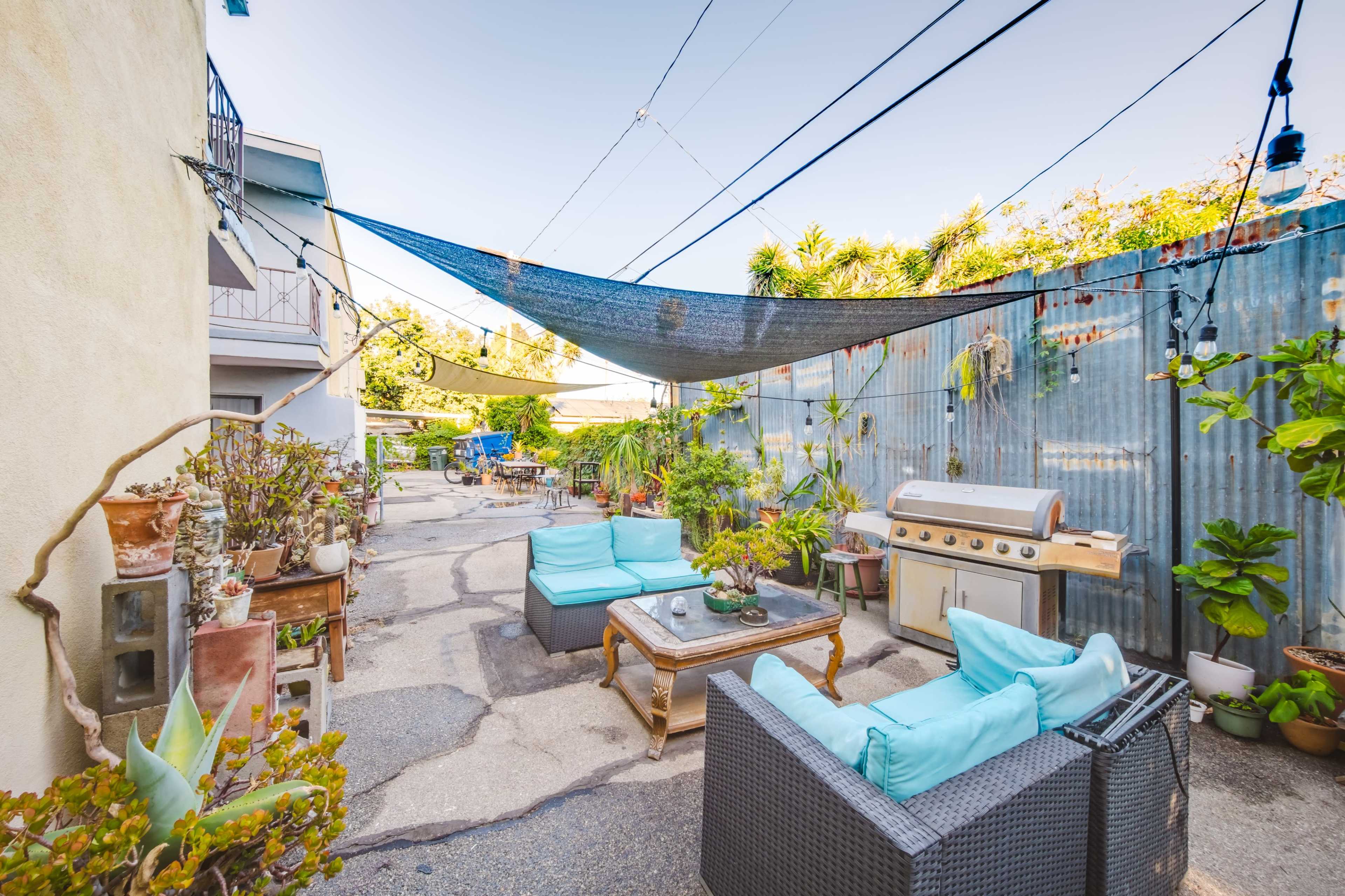 The scene features an outdoor space with comfortable seating, a grill, and various plants, enclosed by a corrugated metal wall and power lines above.