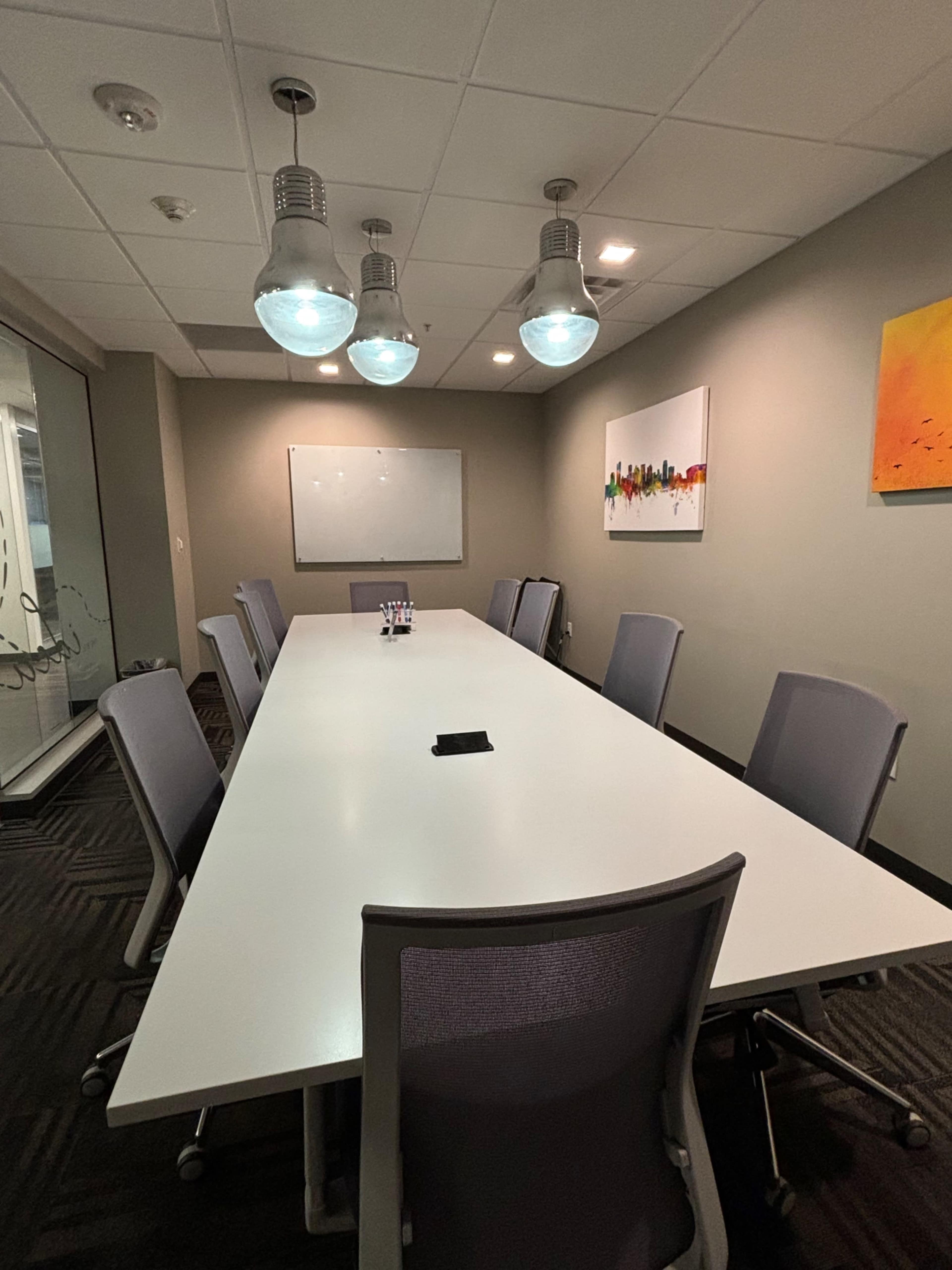 A spacious conference room features a long white table surrounded by eight gray chairs, with large hanging light fixtures and a whiteboard on the wall.