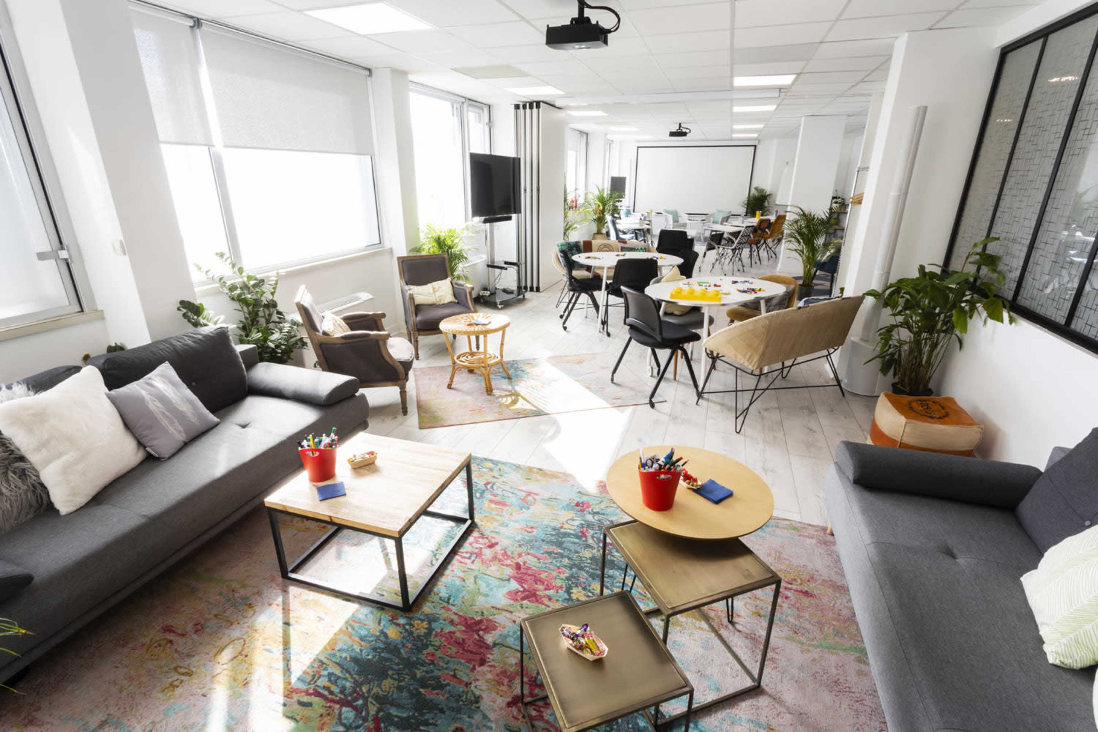 A bright and modern workspace features comfortable seating areas, tables for meetings, and plants throughout the room.