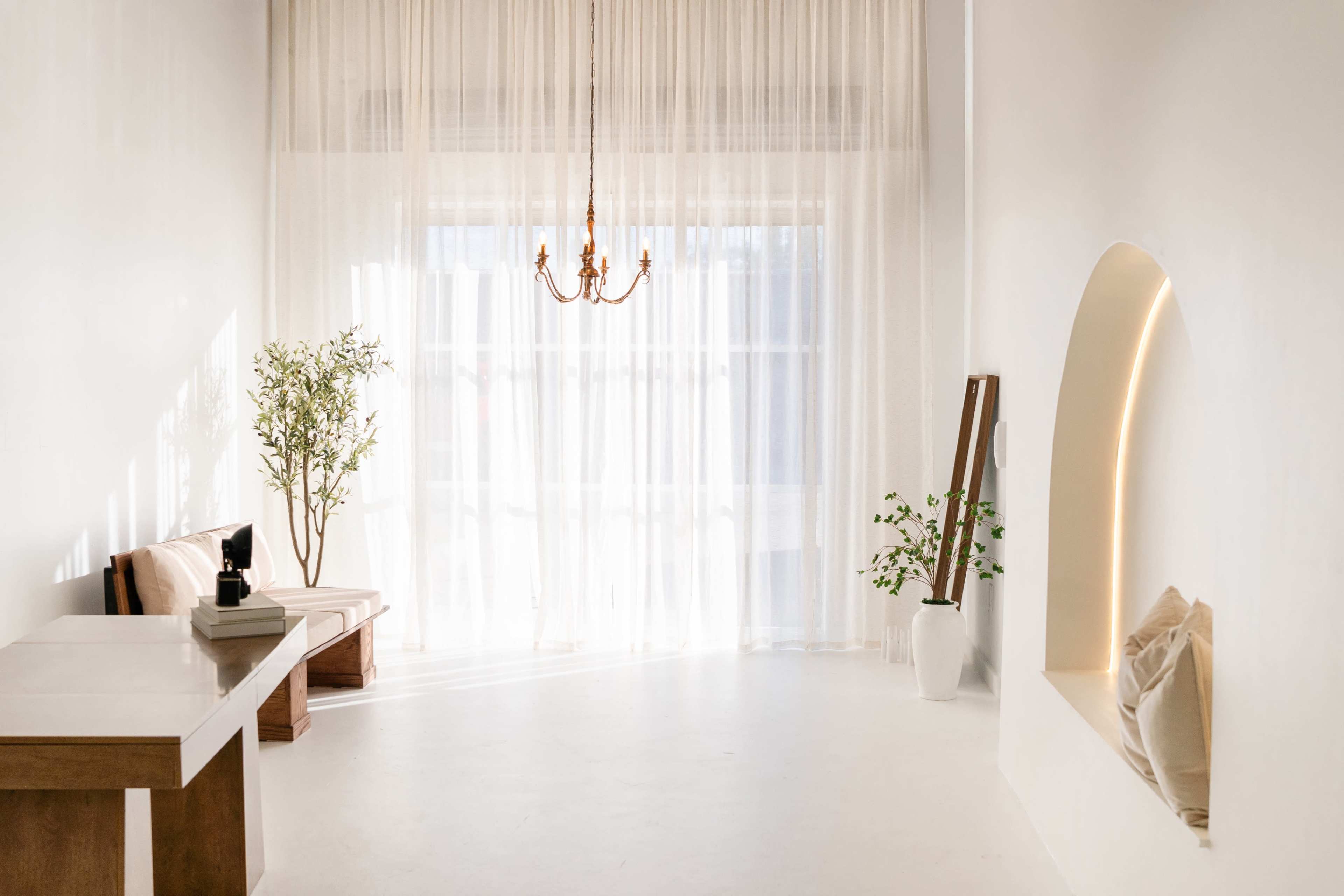 The image shows a minimalist, well-lit room with a chandelier, sheer curtains, a small table, and plants.