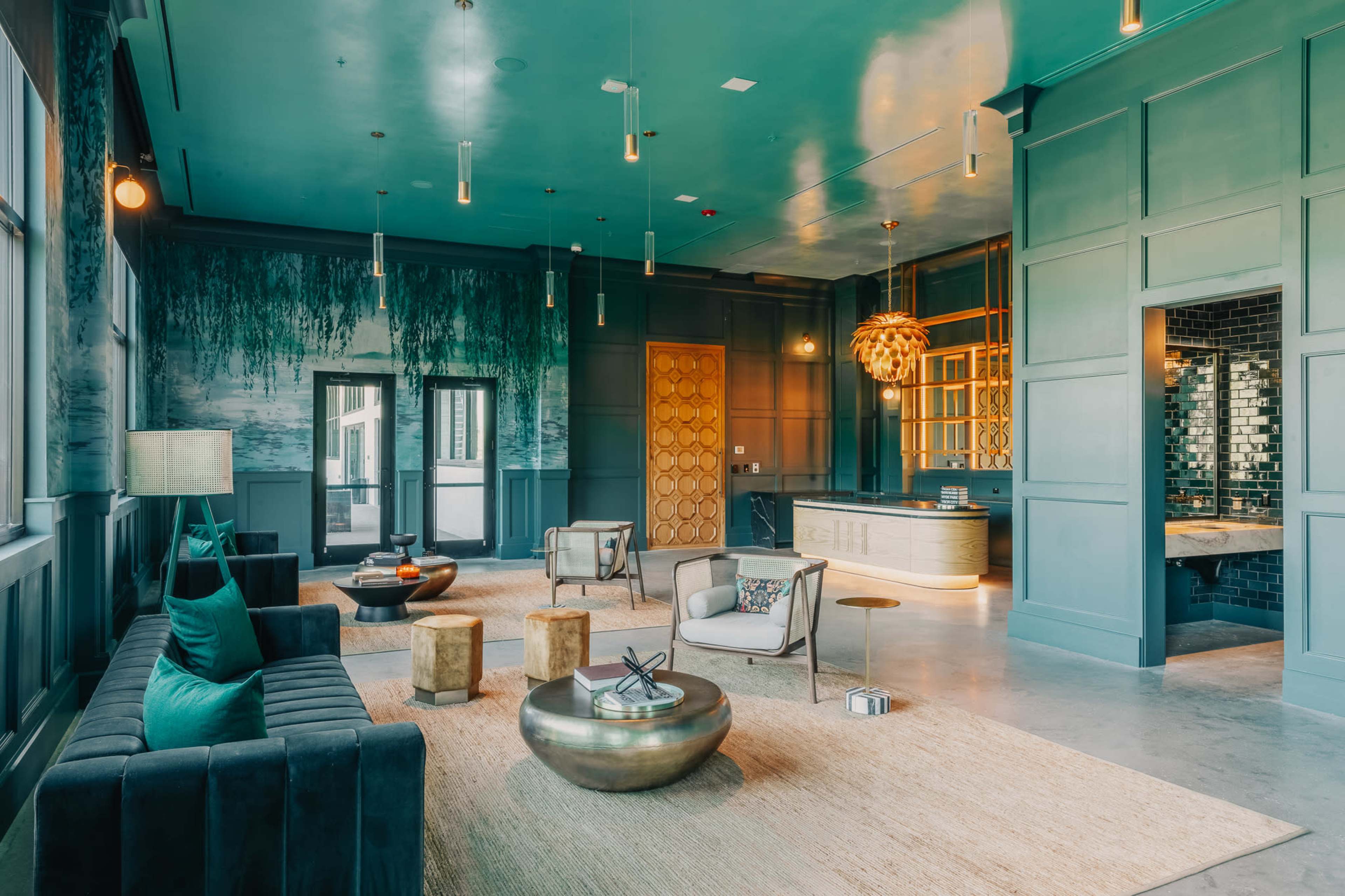 The image shows a modern lounge area with teal walls, stylish seating, and a kitchen area, featuring a mixture of lighting fixtures and decorative elements.