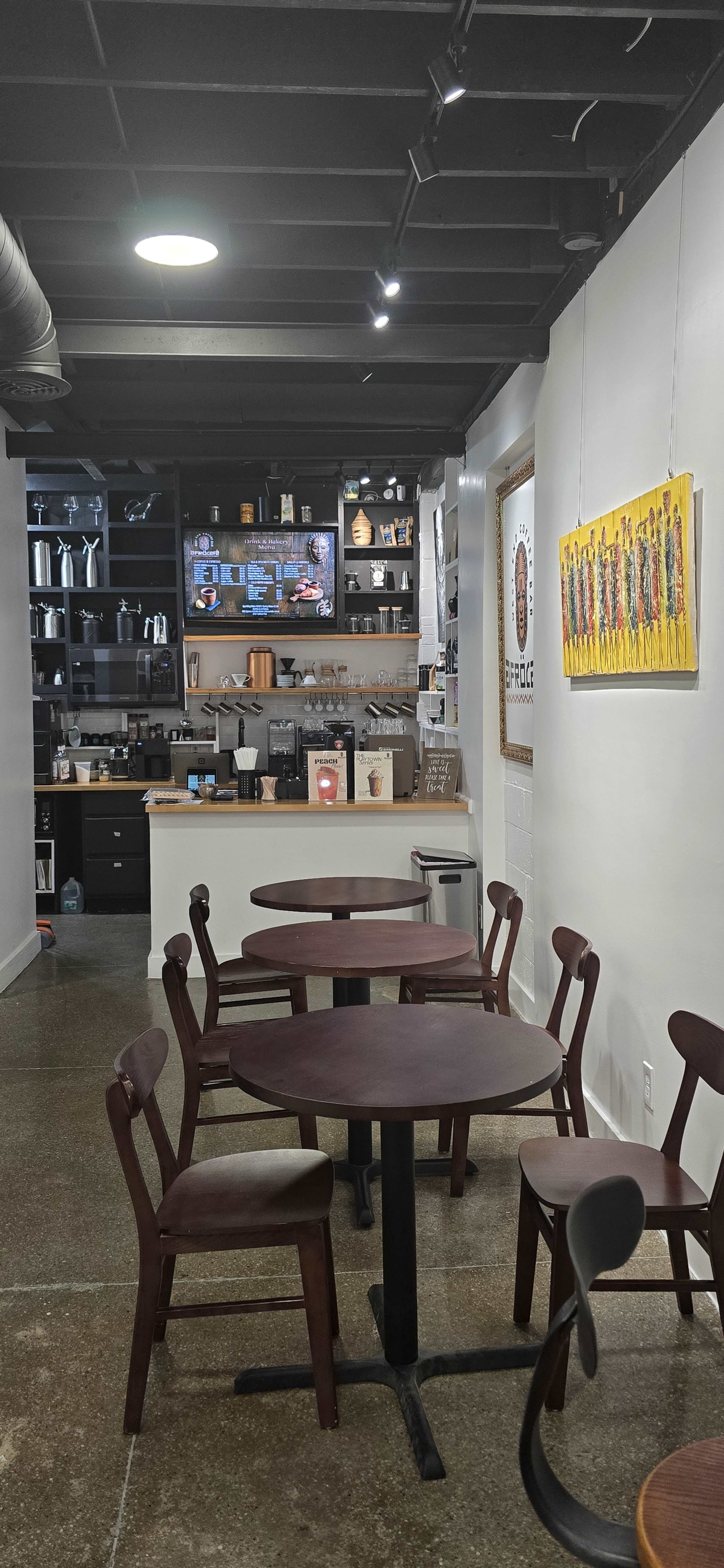 The image shows a cafe interior with several tables and chairs arranged in front of a service counter featuring coffee equipment and a television mounted on the wall.