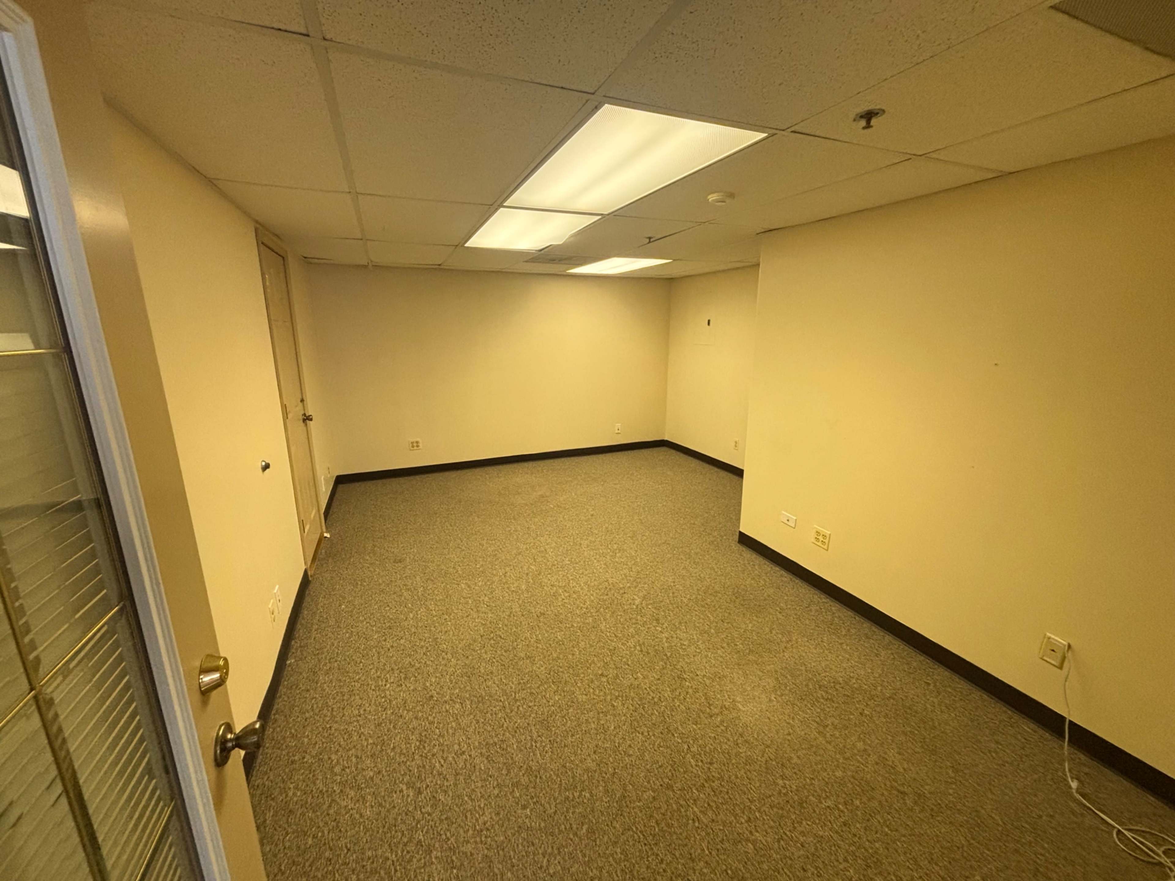 The image shows a vacant office room with beige walls, gray carpeting, and a single door leading to another space.