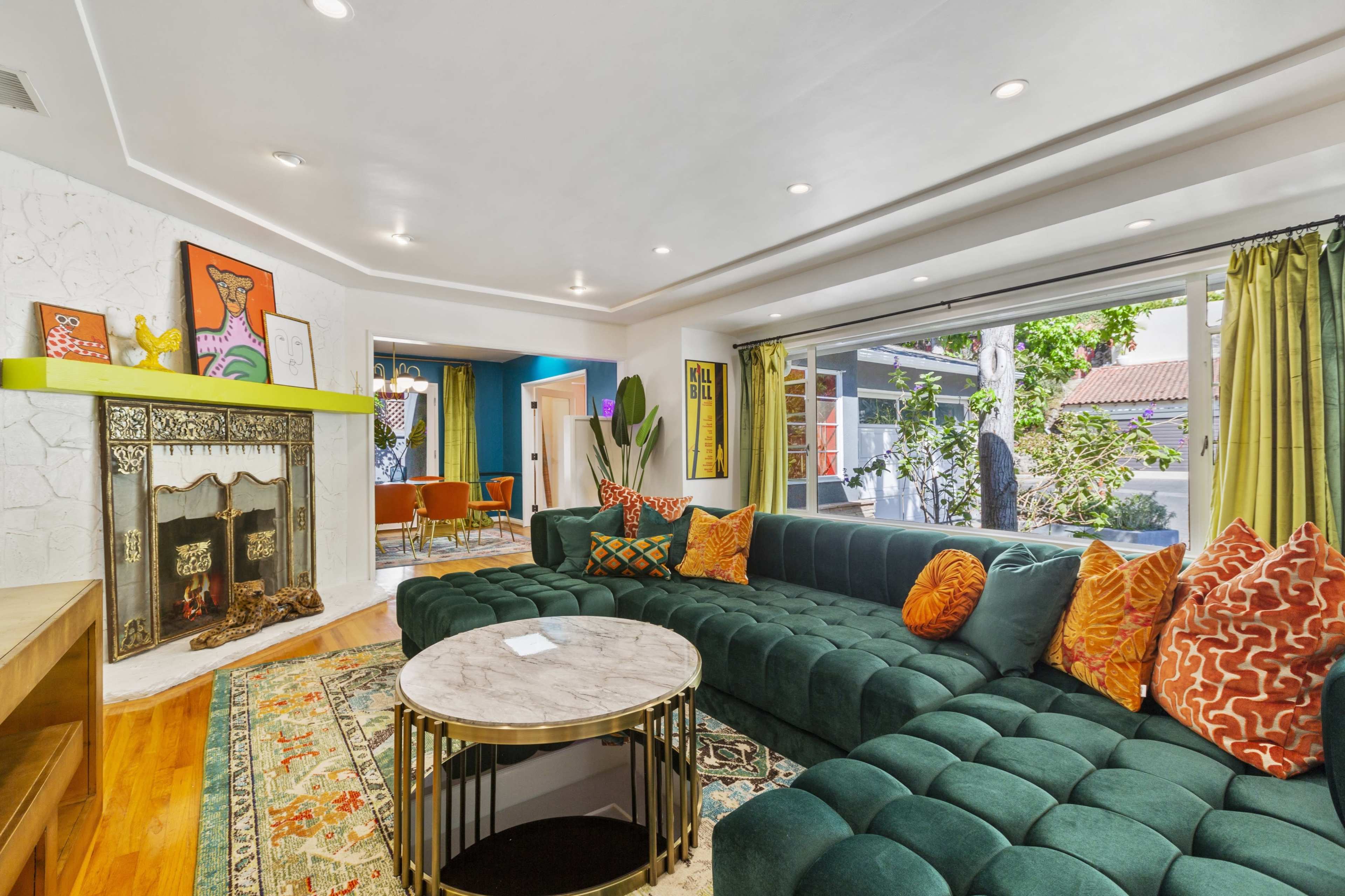 The living room features a large green sectional couch, a marble coffee table, and a decorative fireplace with colorful artwork in the background.