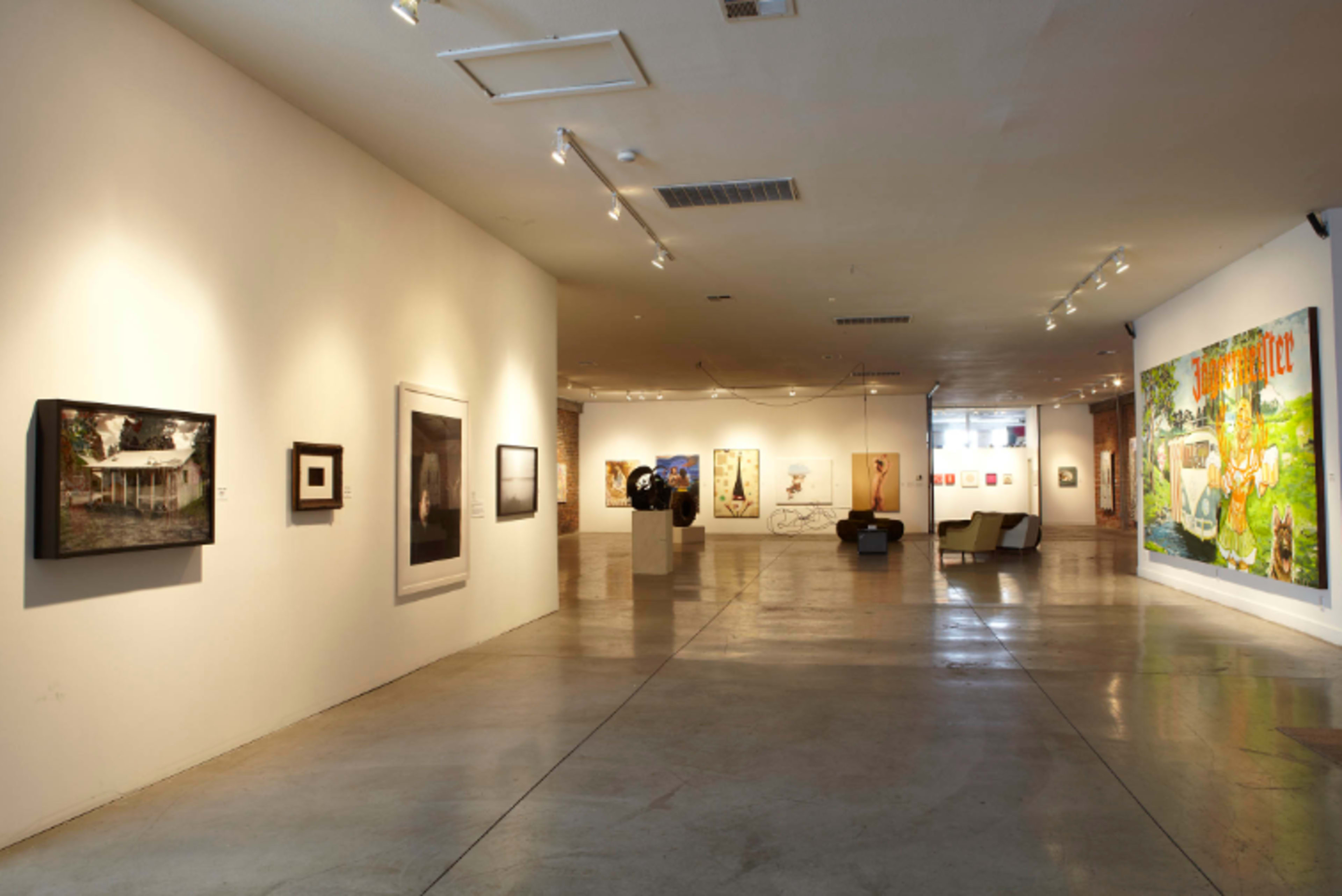 A spacious art gallery featuring a variety of framed artworks on white walls and a seating area in the background.