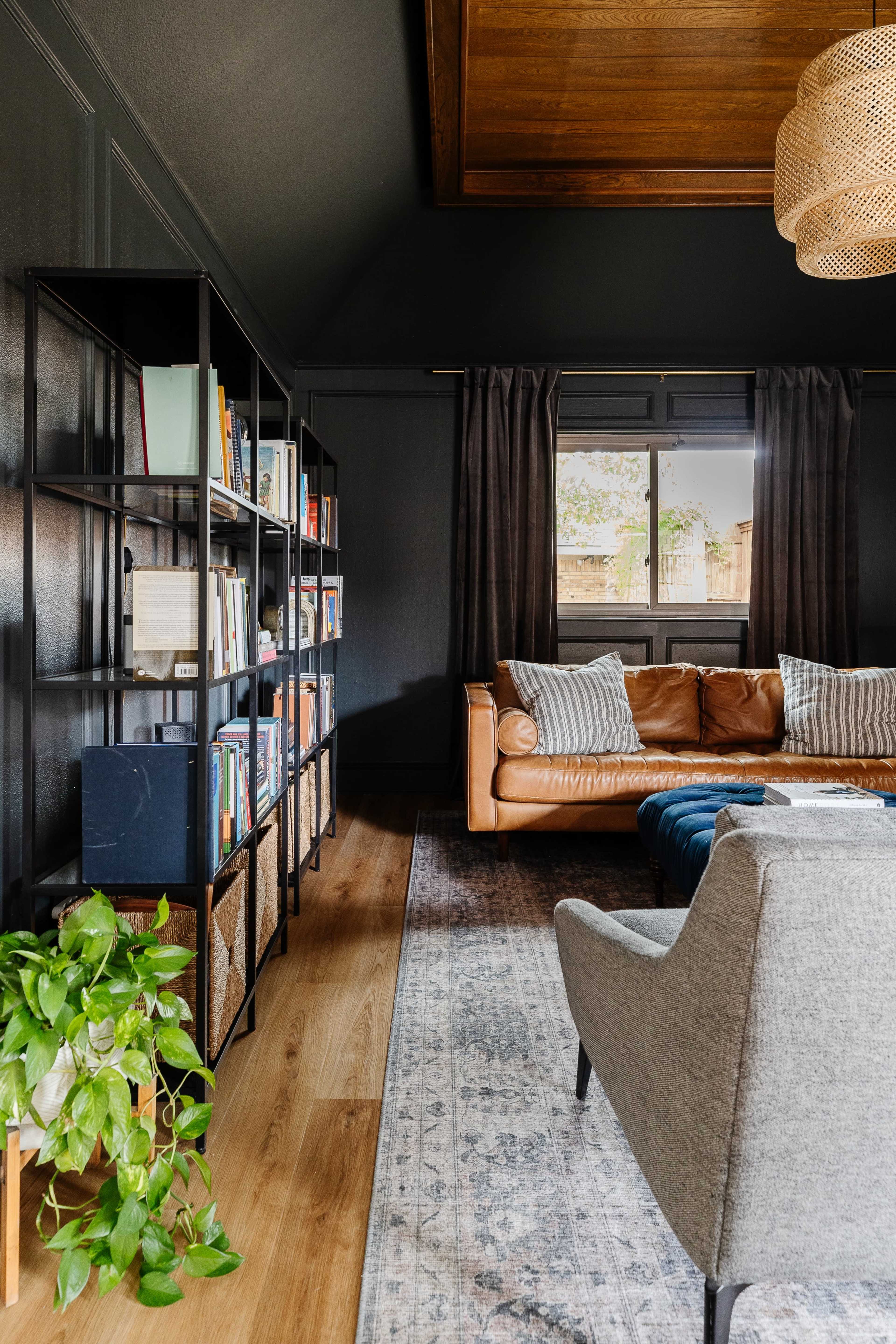 A modern living room features a leather sofa, a gray armchair, a tall bookshelf with books, and a large window with dark curtains.