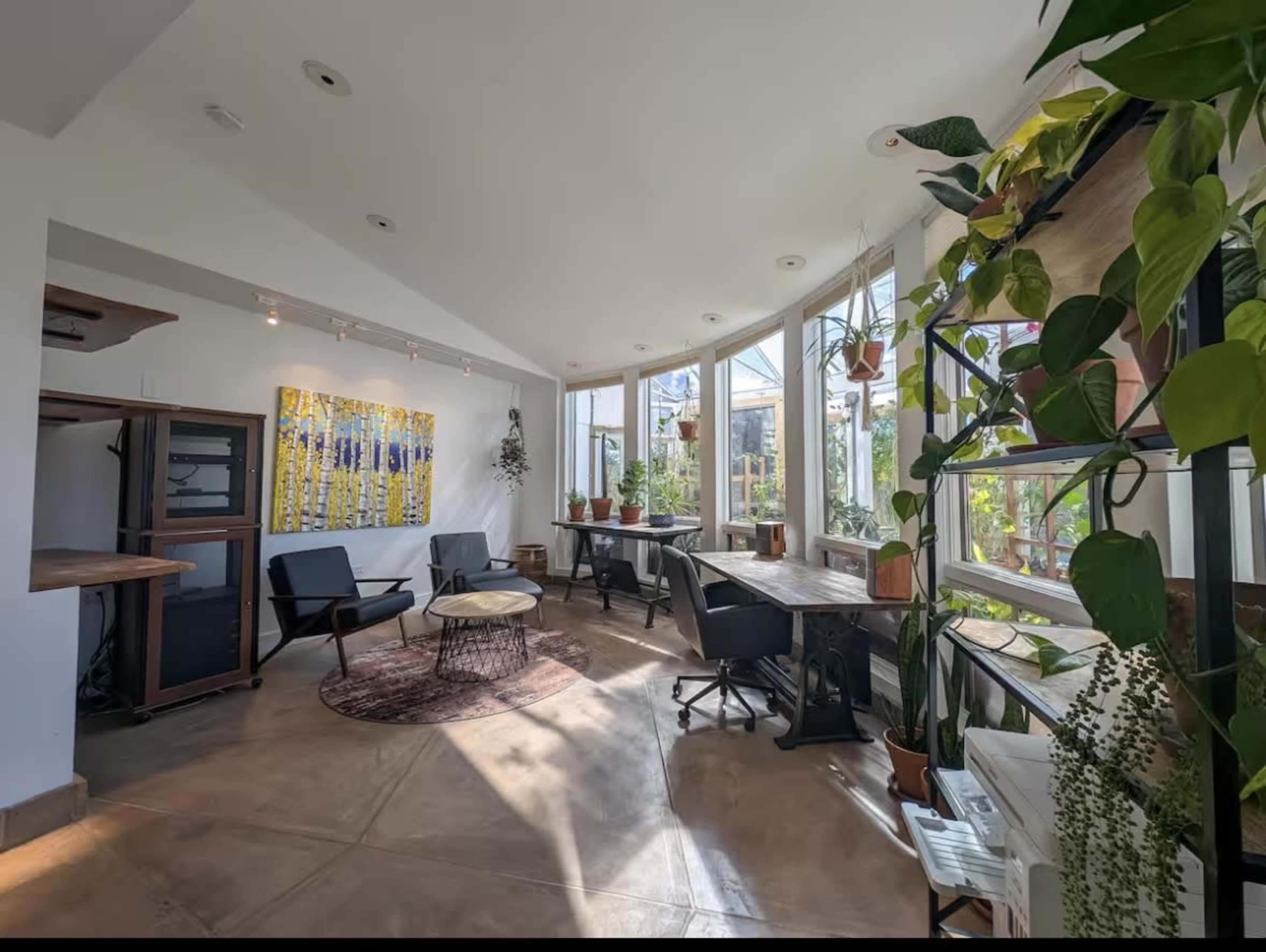 The image shows a bright, modern workspace with large windows, wooden desks, and plants, creating an open and airy atmosphere.