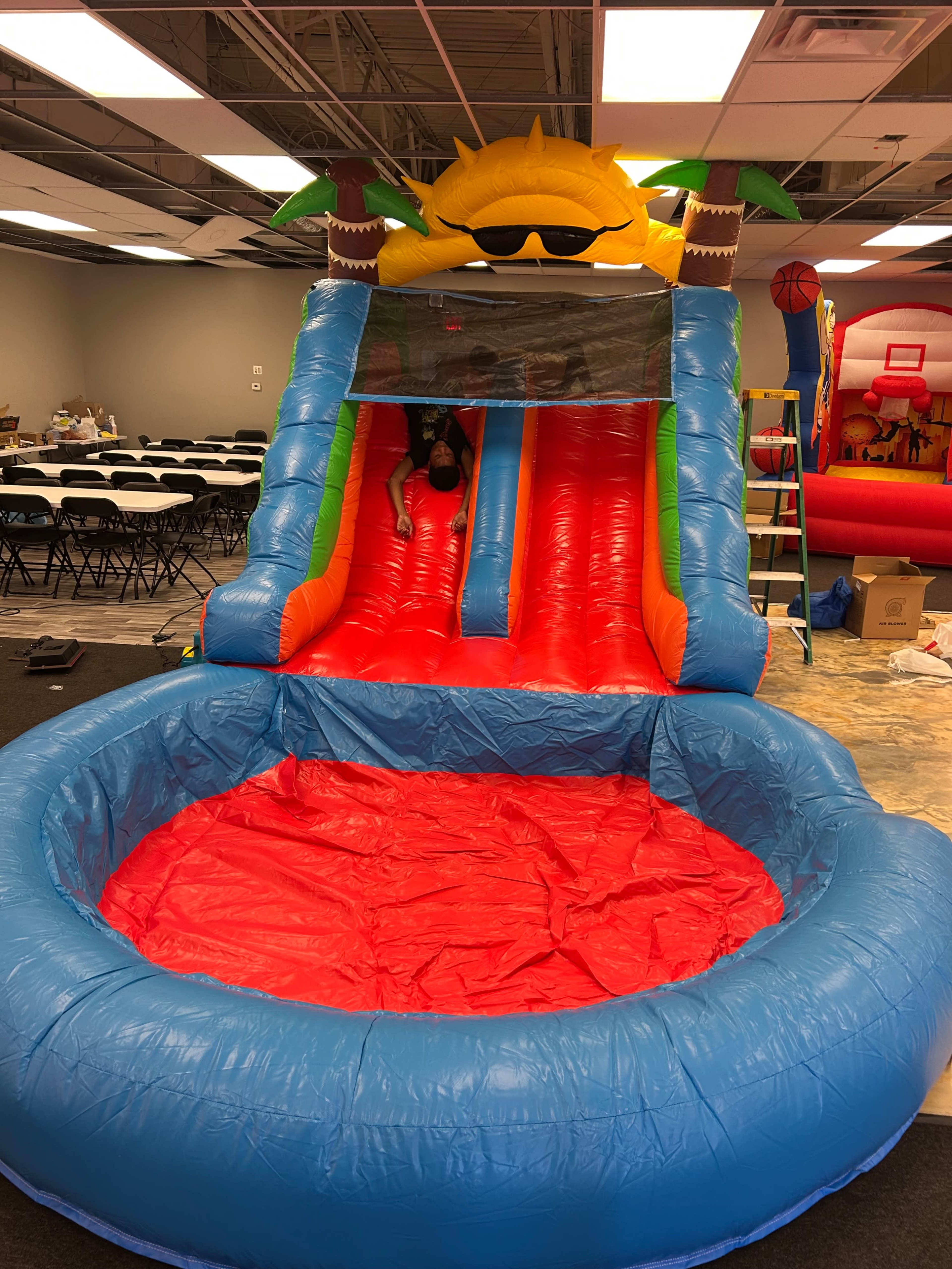 The image shows an inflatable water slide with a bright, colorful design featuring a sun with sunglasses at the top and a red landing area.