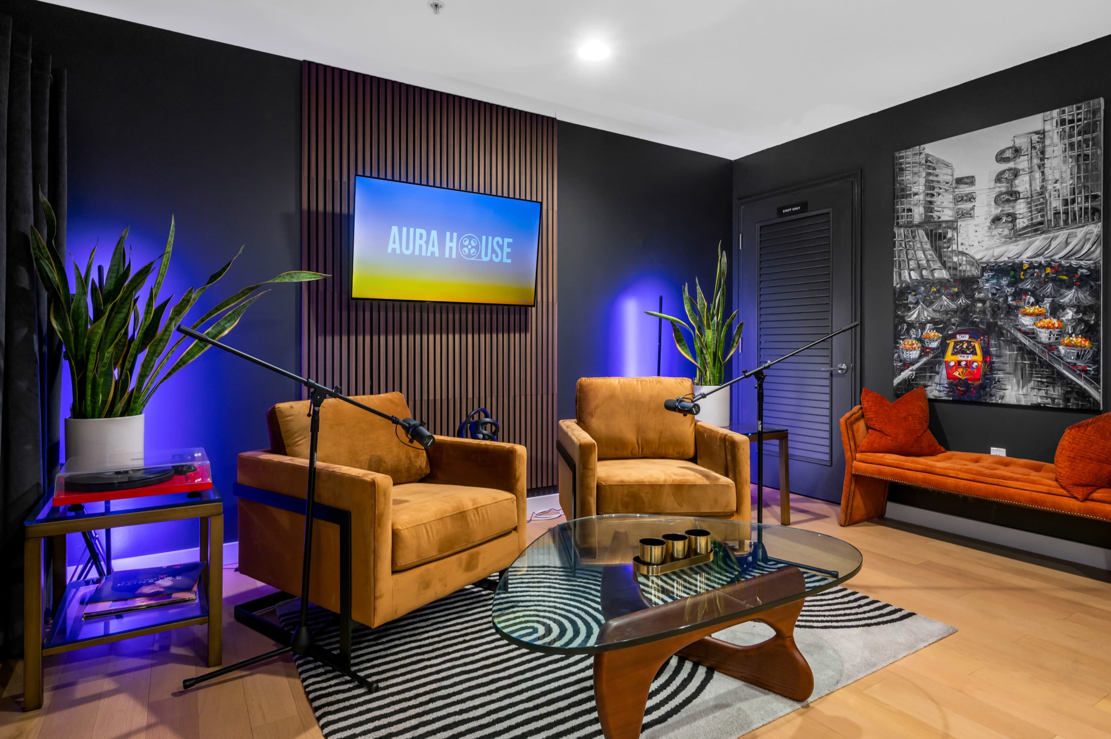 A modern room with two orange chairs, a glass coffee table, a wall-mounted TV, and a large artwork on one side, accompanied by green plants and decorative lighting.