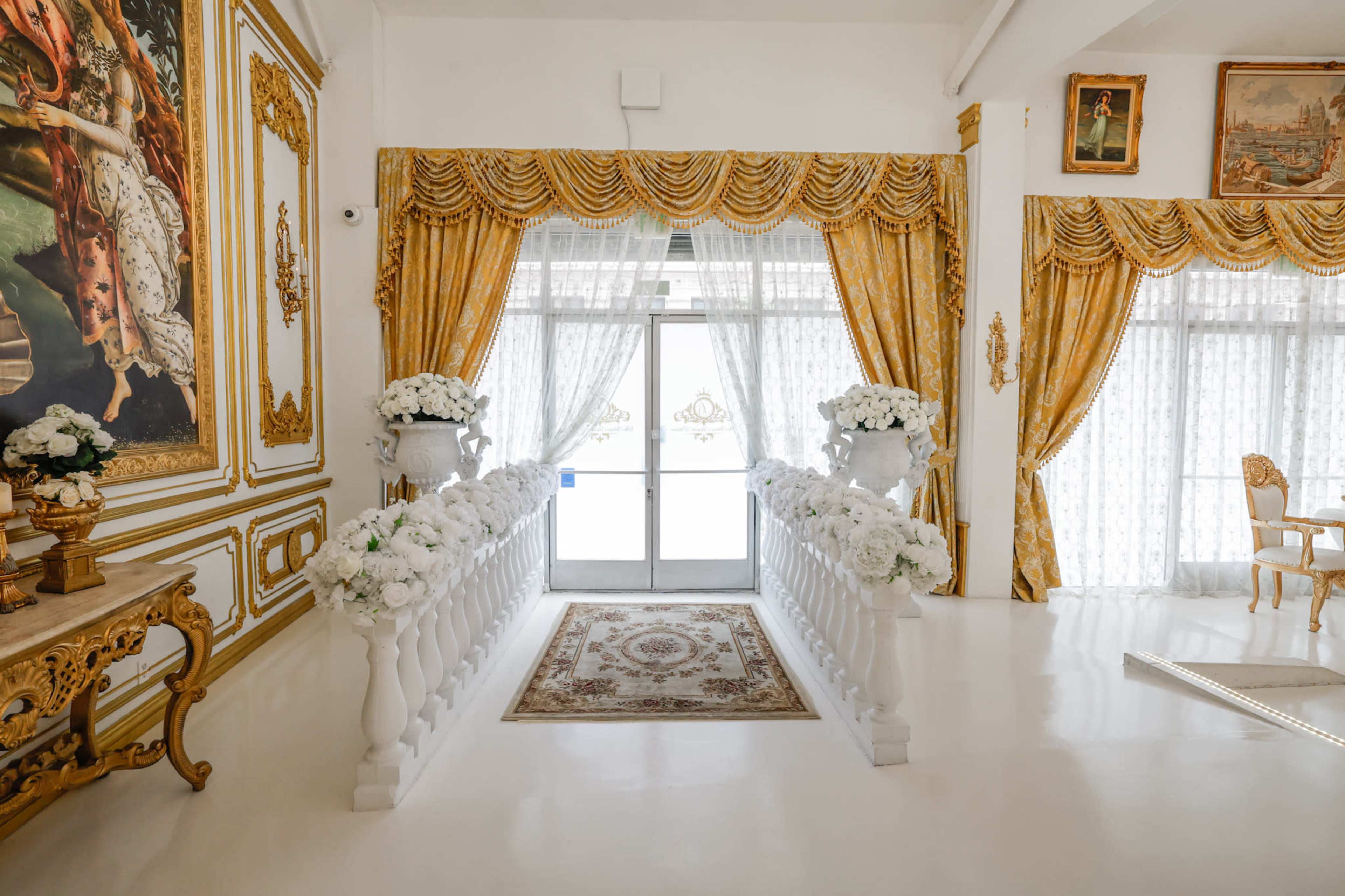 The image shows an elegantly decorated entrance area with white walls, gold drapes, floral arrangements, and a carpet runner leading to a glass door.