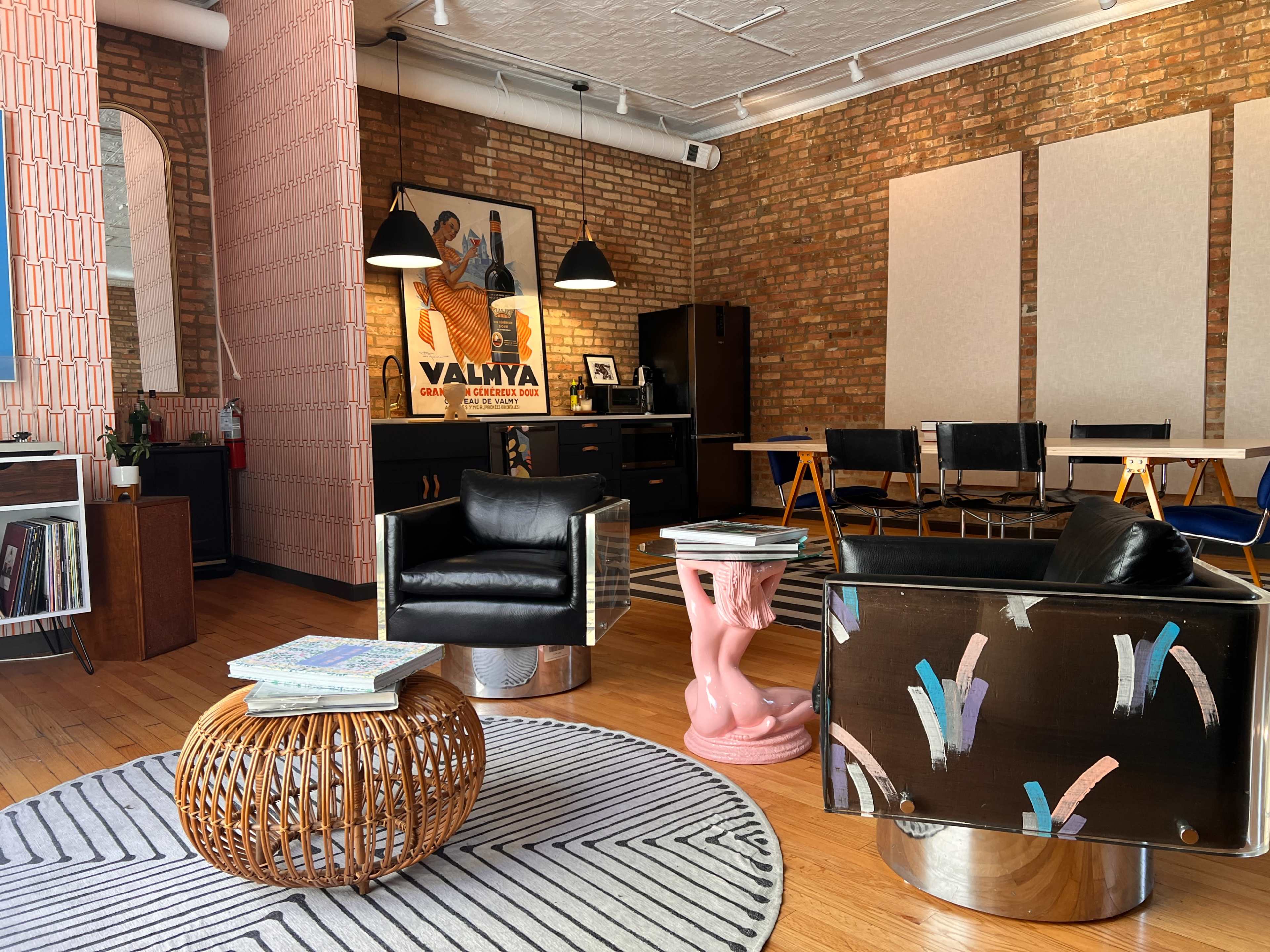 The image shows a modern interior space featuring a mix of seating options, including black chairs and a pink sculpture table, with brick walls and decorative accents.
