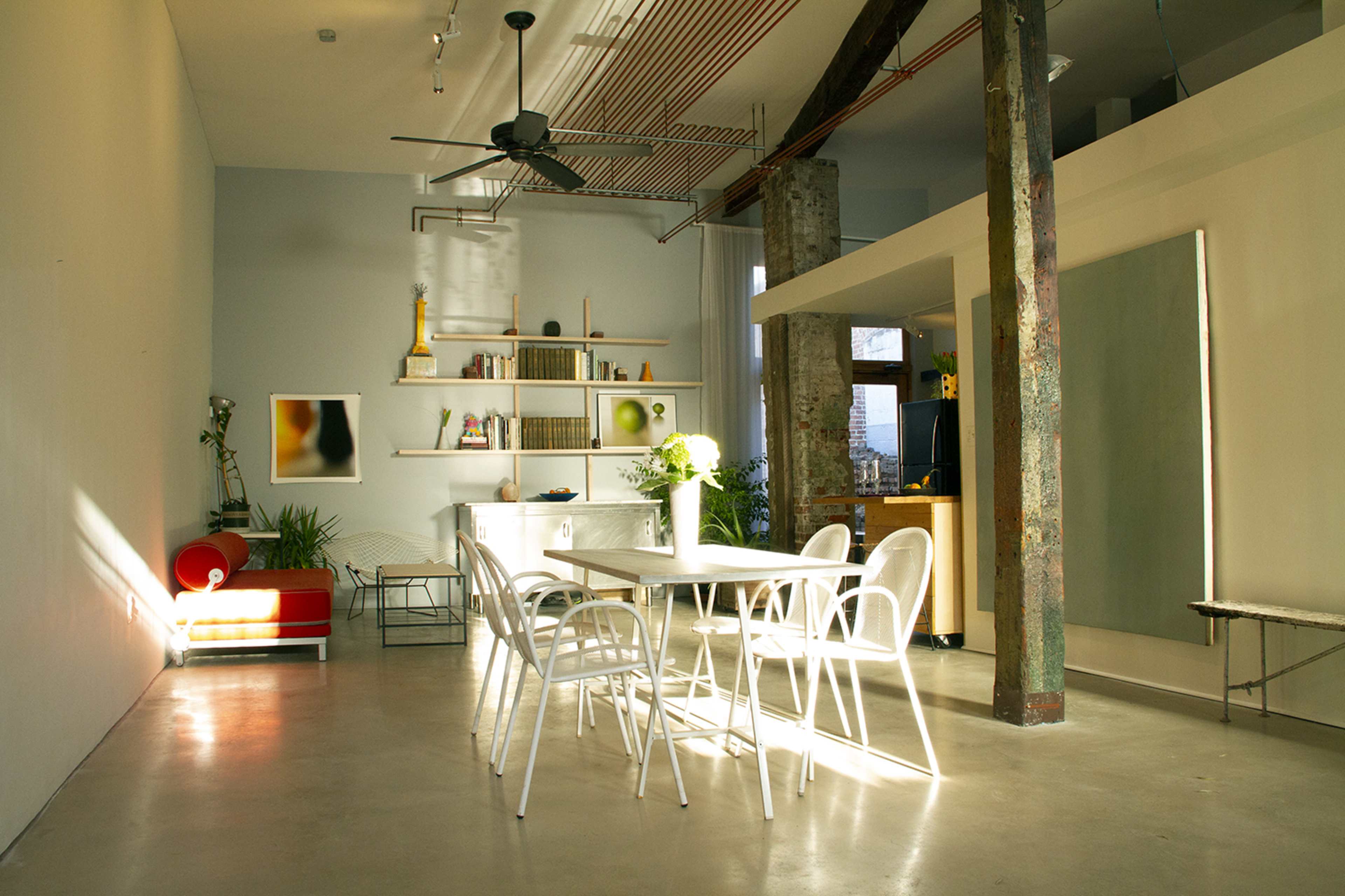 A modern, minimalist dining area with a large table surrounded by white chairs, a kitchenette in the background, and natural light streaming through the windows.