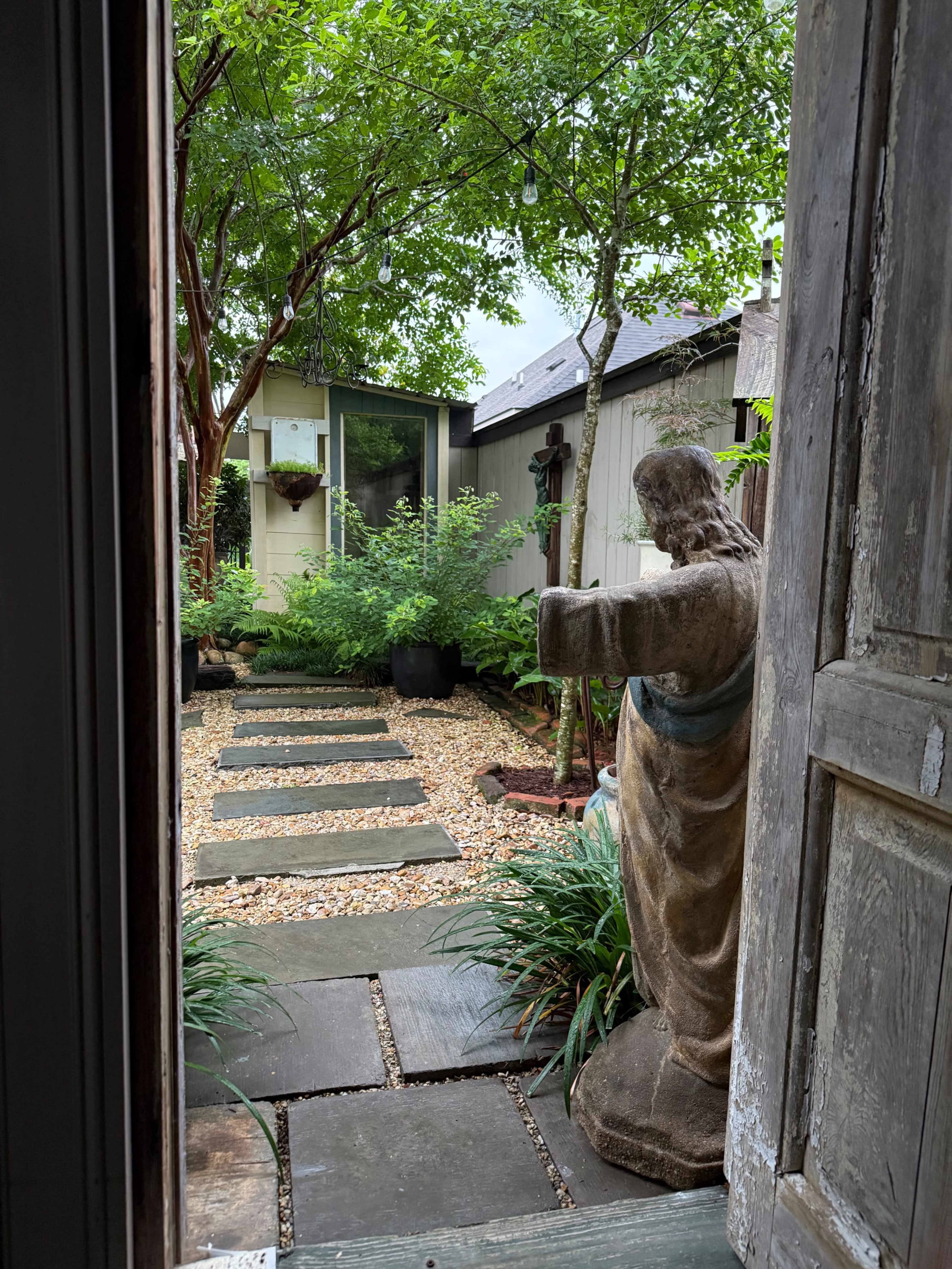 A weathered statue stands at the entrance of a gravel pathway leading to a landscaped garden with greenery and a small structure in the background.