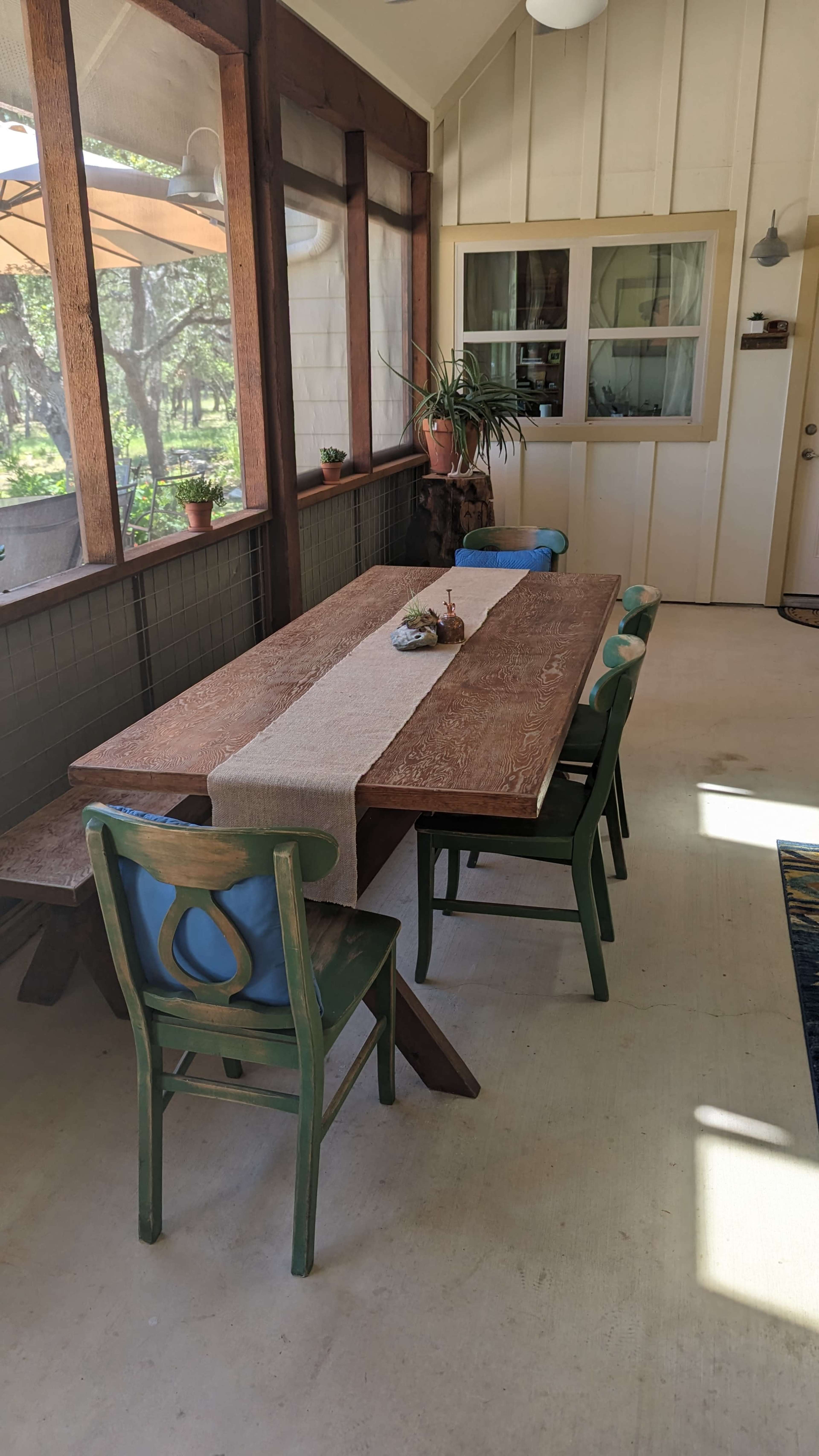 A wooden dining table with a runner sits amid green chairs in a sunlit room lined with windows overlooking a garden.