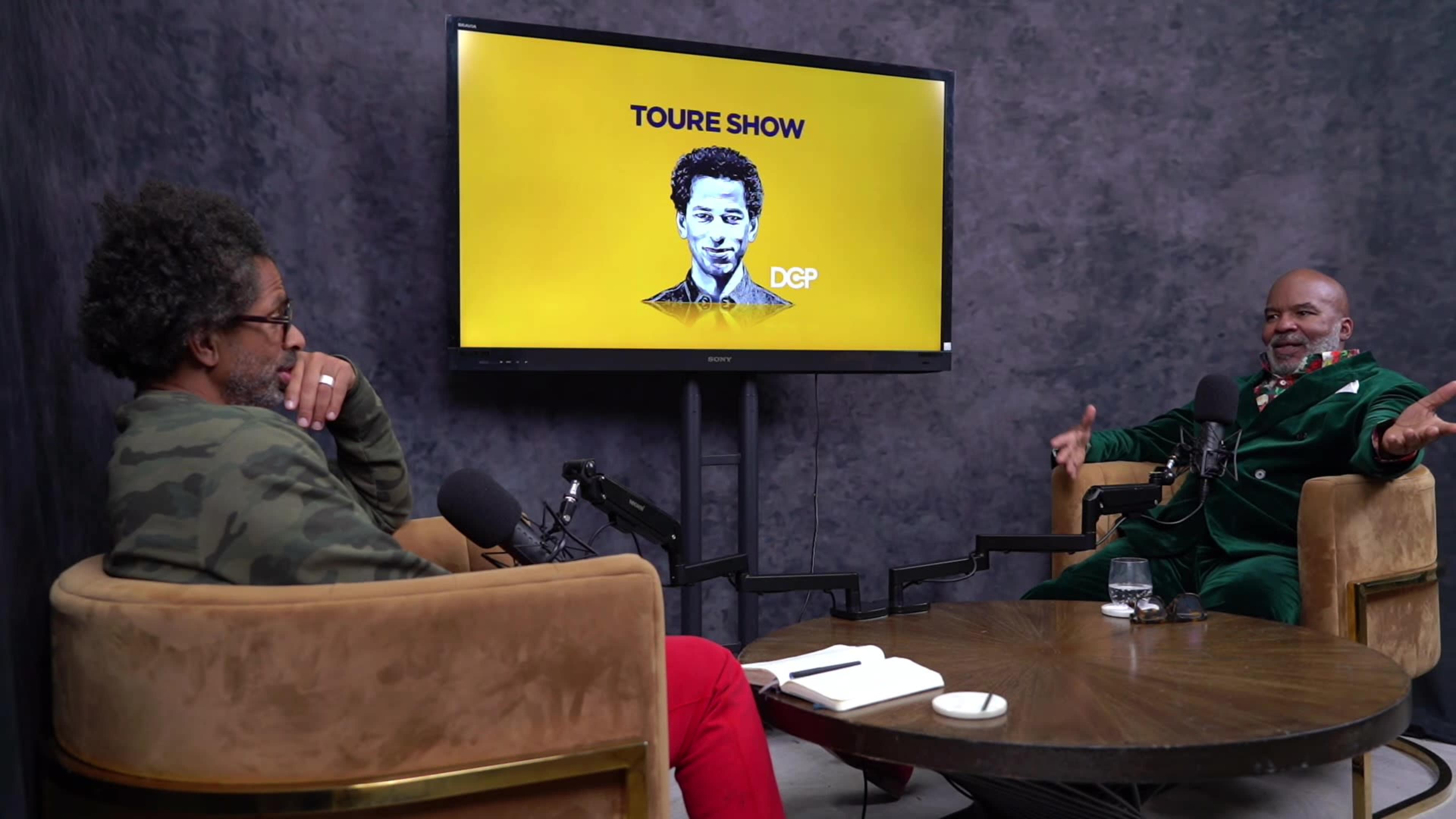 A podcast set features two men sitting in chairs in front of a large screen displaying the title "TOURE SHOW" and an image of a man.