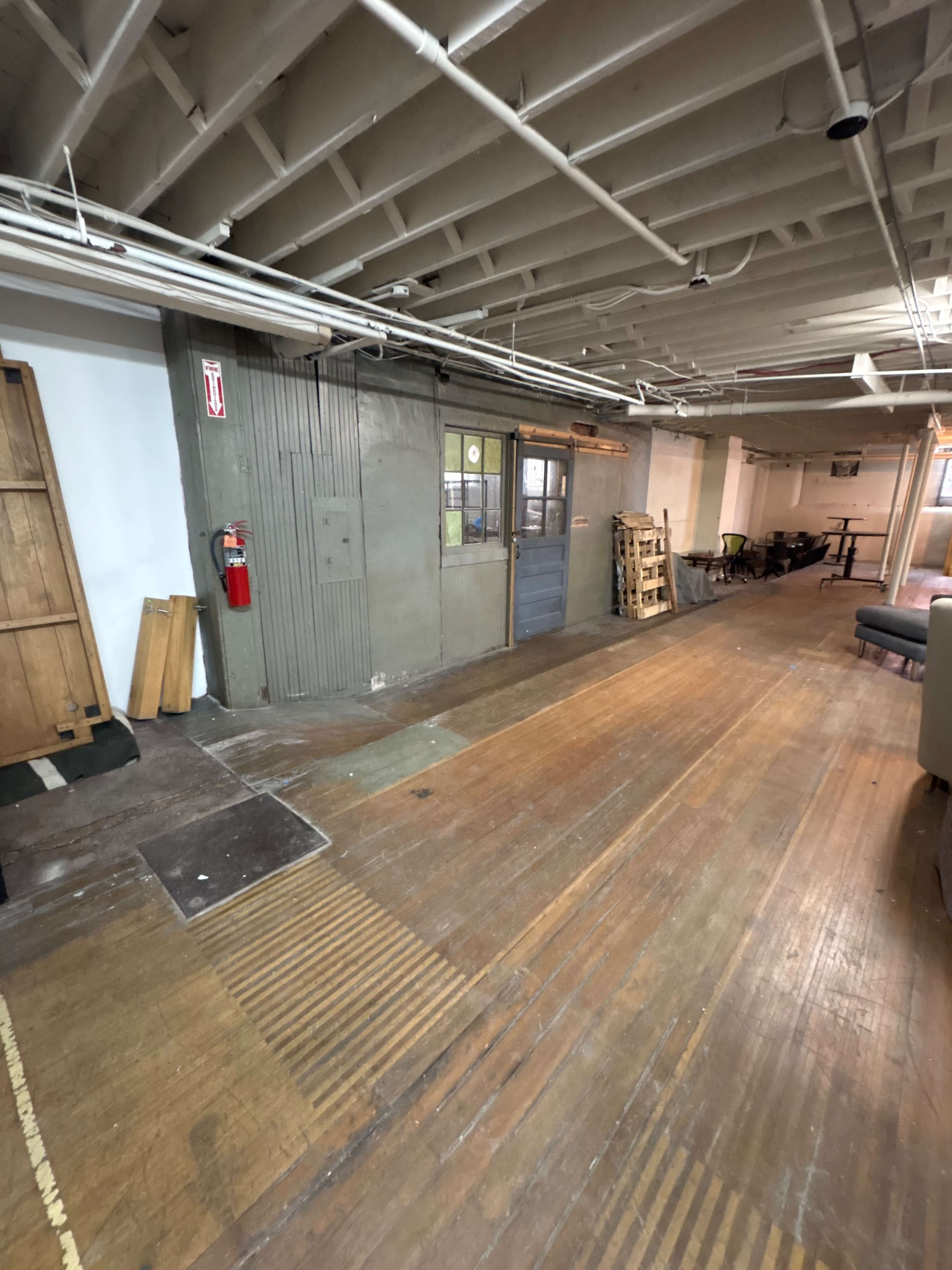 The image shows a warehouse-style interior with a wooden floor, exposed pipes, and a gray metal door at the far end.