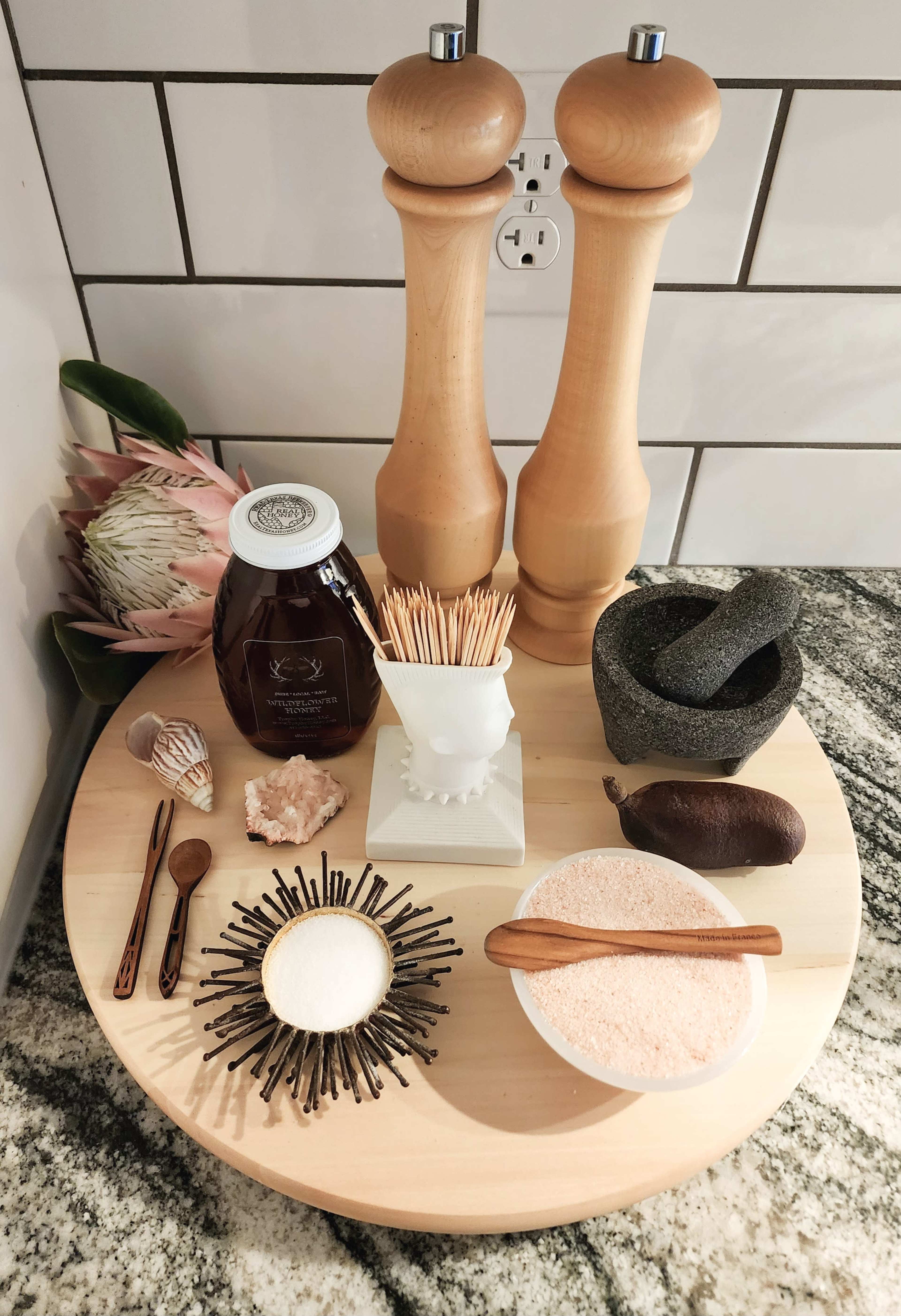 A wooden round platter displays two pepper mills, honey in a jar, various utensils, a mortar and pestle, decorative shells, and bowls of salt and pink Himalayan salt.