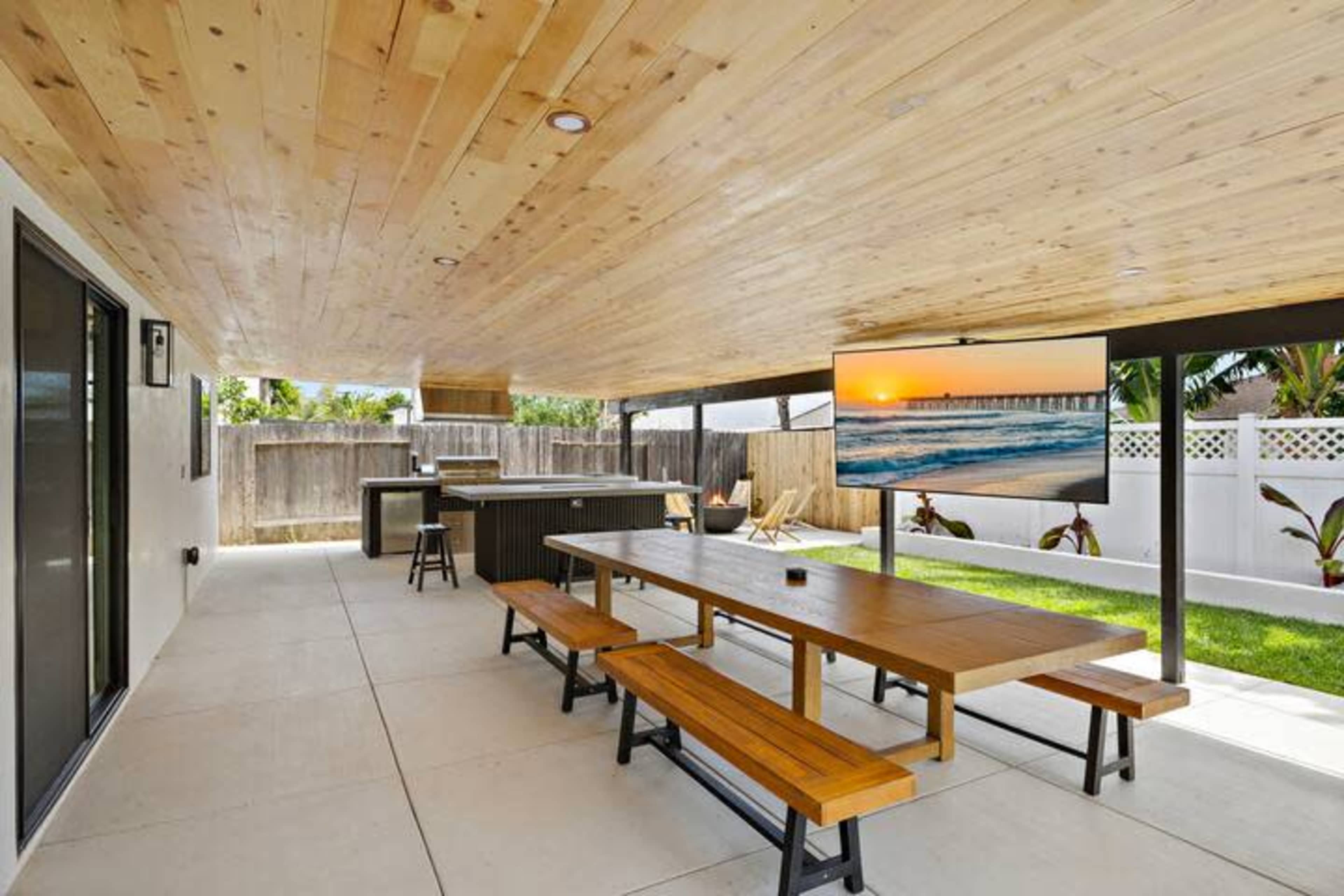 The image shows a spacious outdoor patio area featuring a wooden ceiling, a large dining table, an outdoor kitchen, and a TV mounted on the wall, surrounded by greenery and a fenced yard.