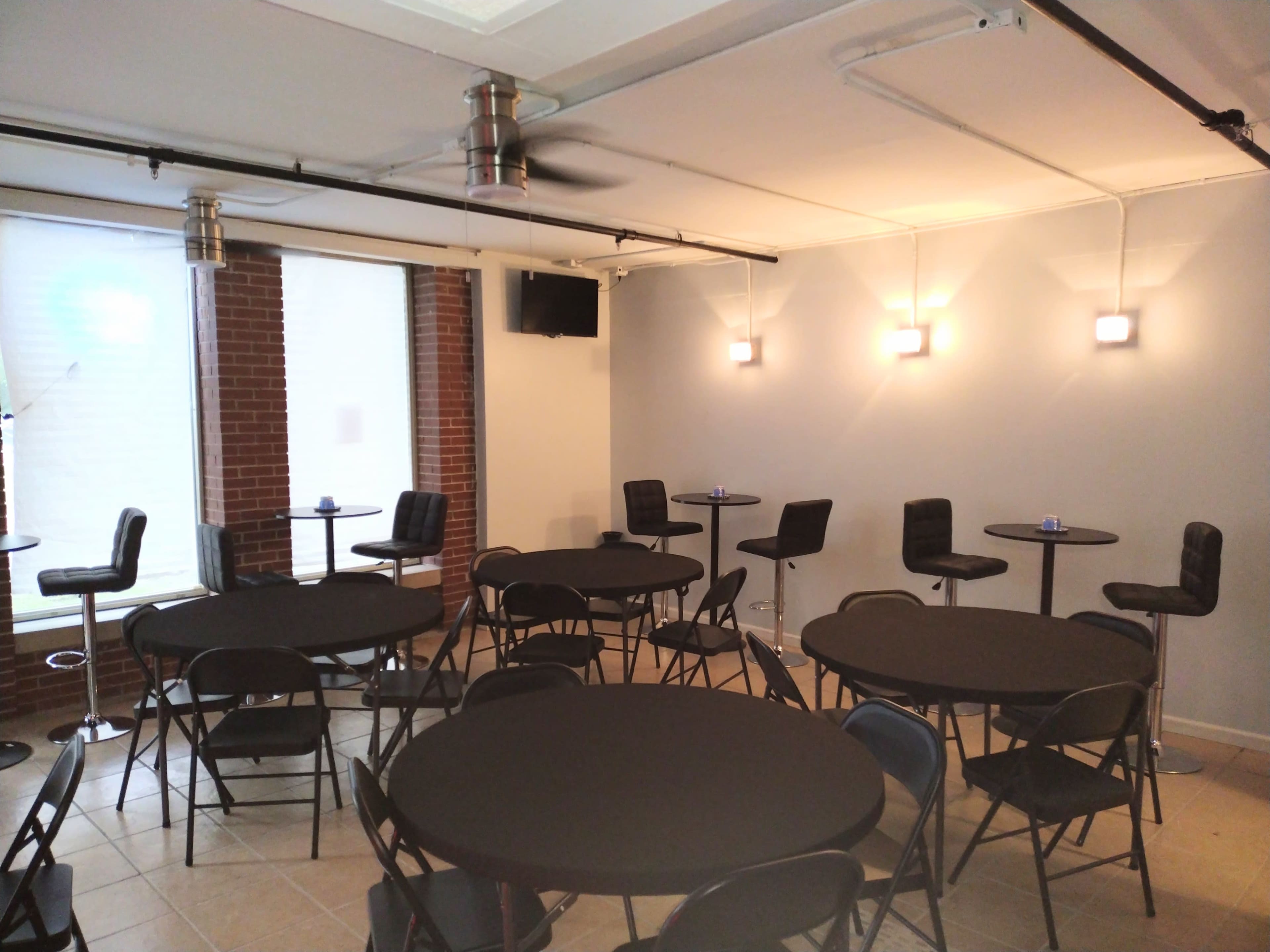 The image shows a spacious room with several circular tables covered in black tablecloths and high-top chairs arranged around them, illuminated by wall-mounted lights.
