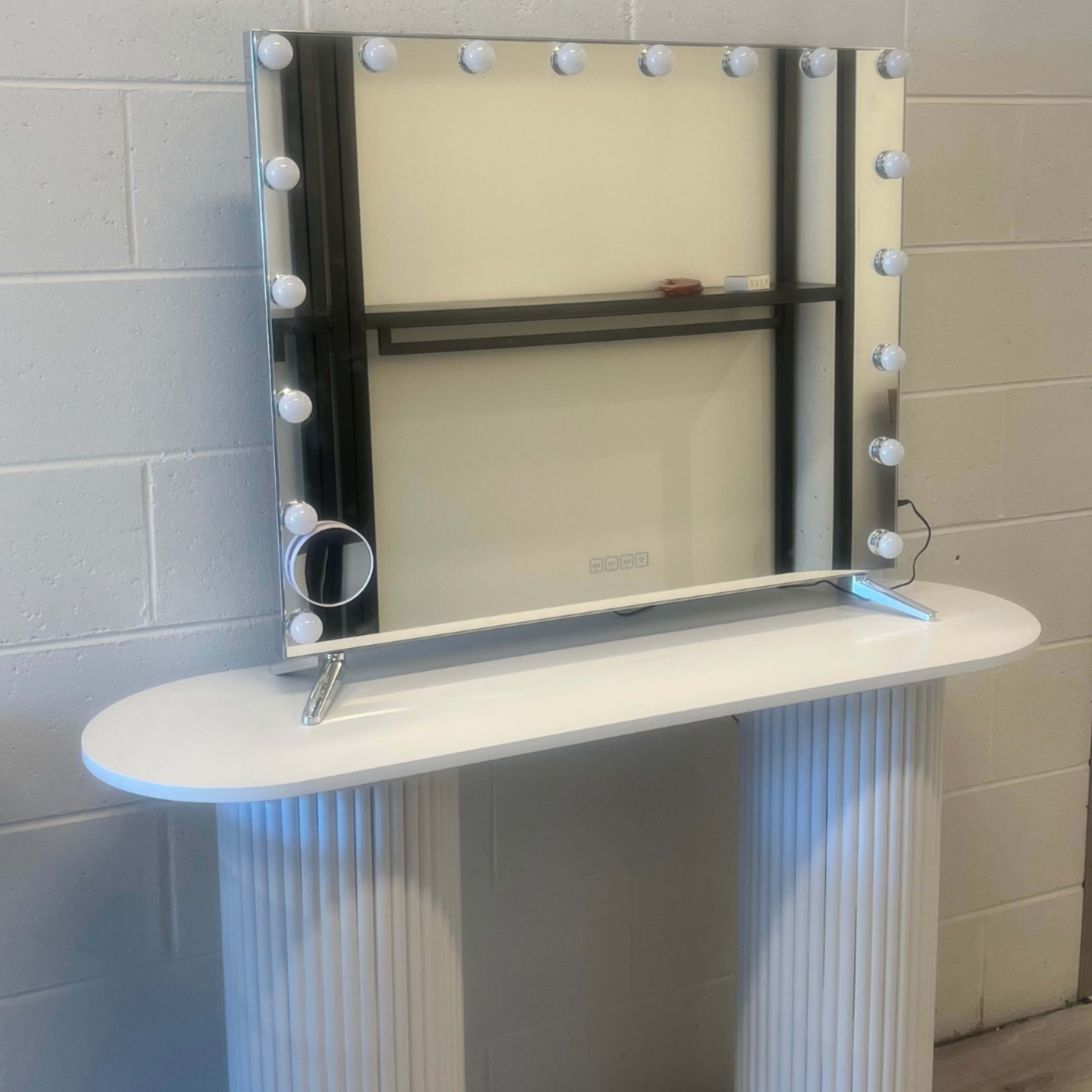 The image shows a vanity mirror with bright bulbs around its edges, mounted above a white console table with fluted columns.