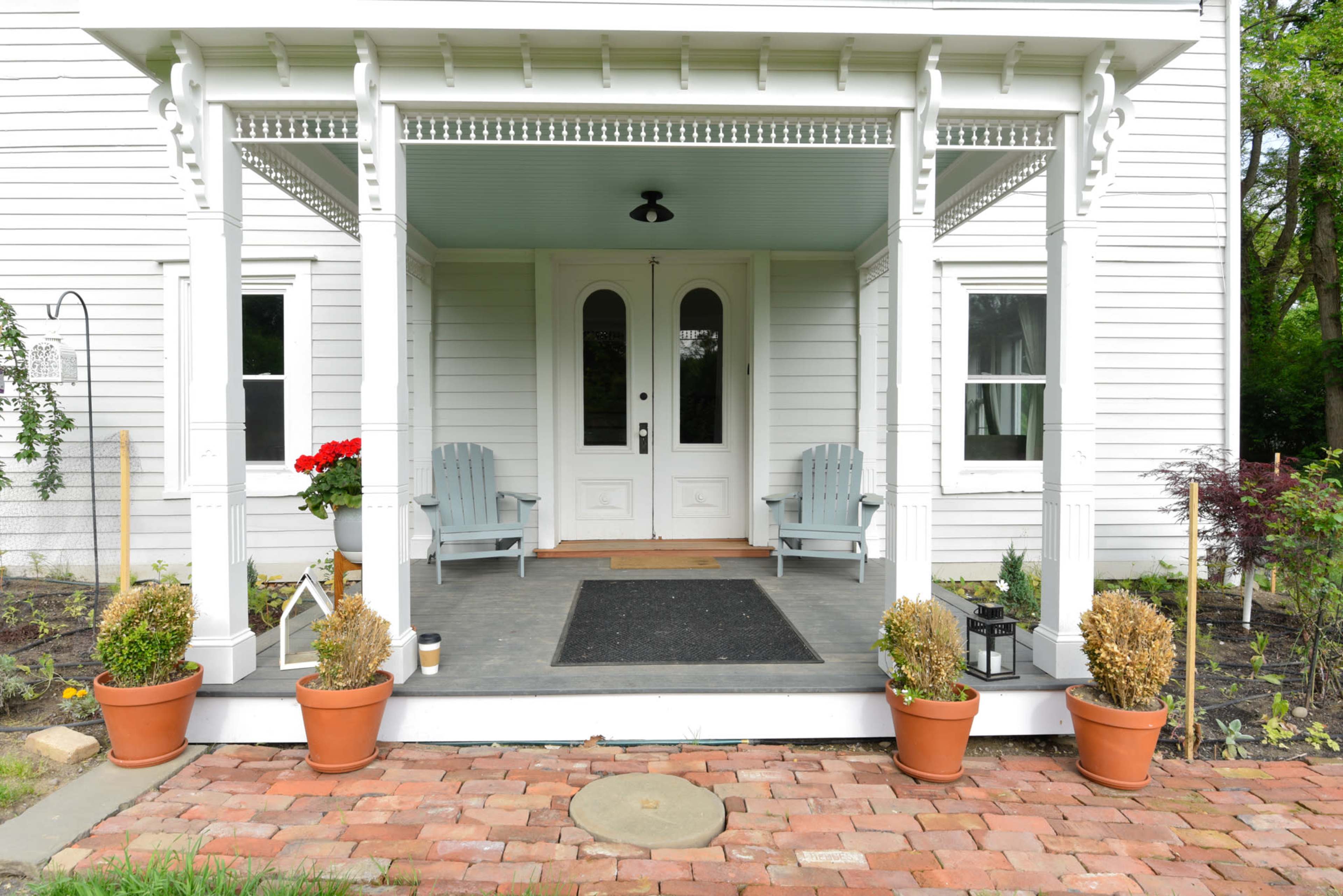 The image shows a covered porch with two light blue chairs and potted plants on either side, leading to a double door entrance of a white house.