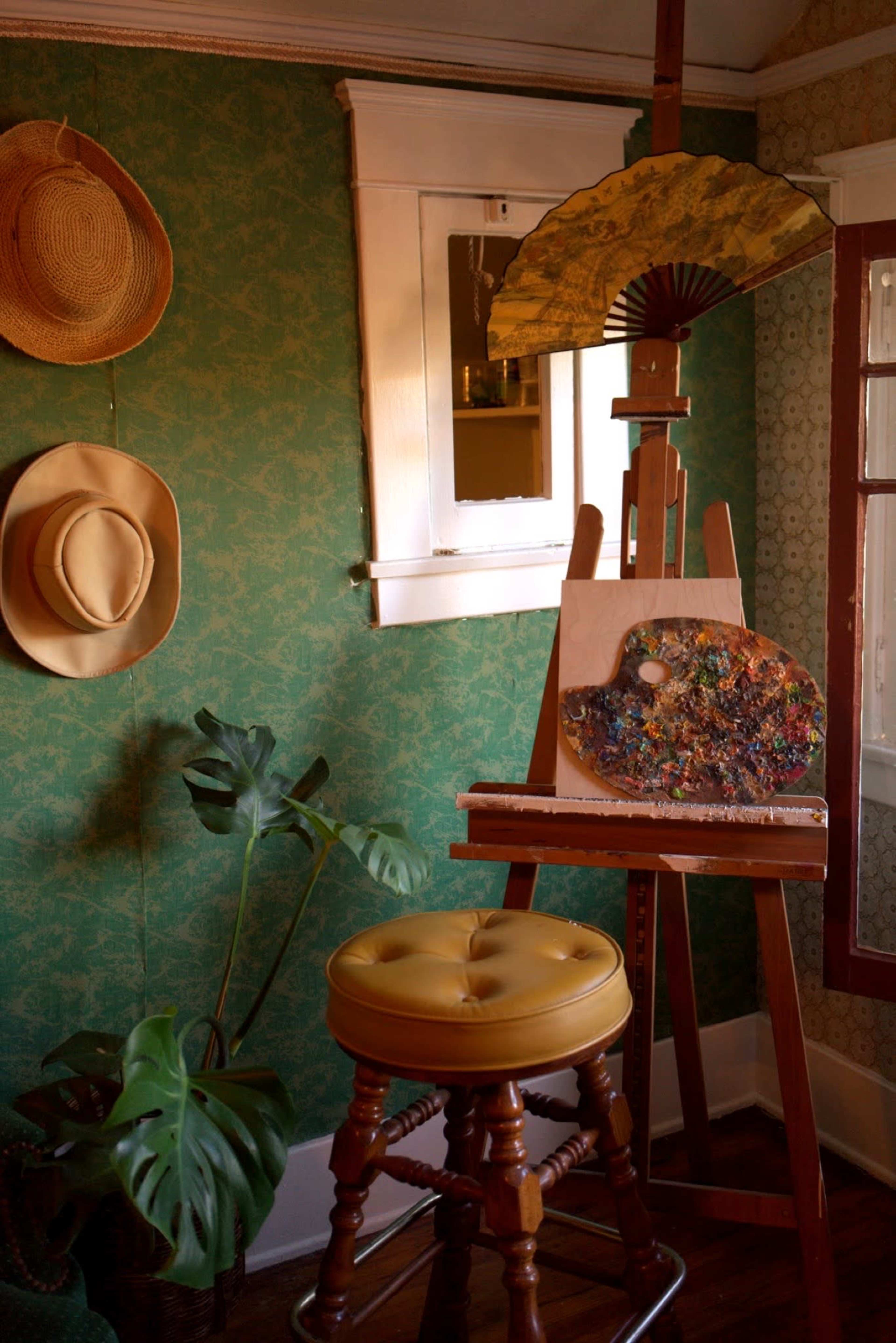 An easel displaying a painted canvas is positioned beside a stool and framed by hats on the wall and a potted plant in a room with patterned wallpaper.