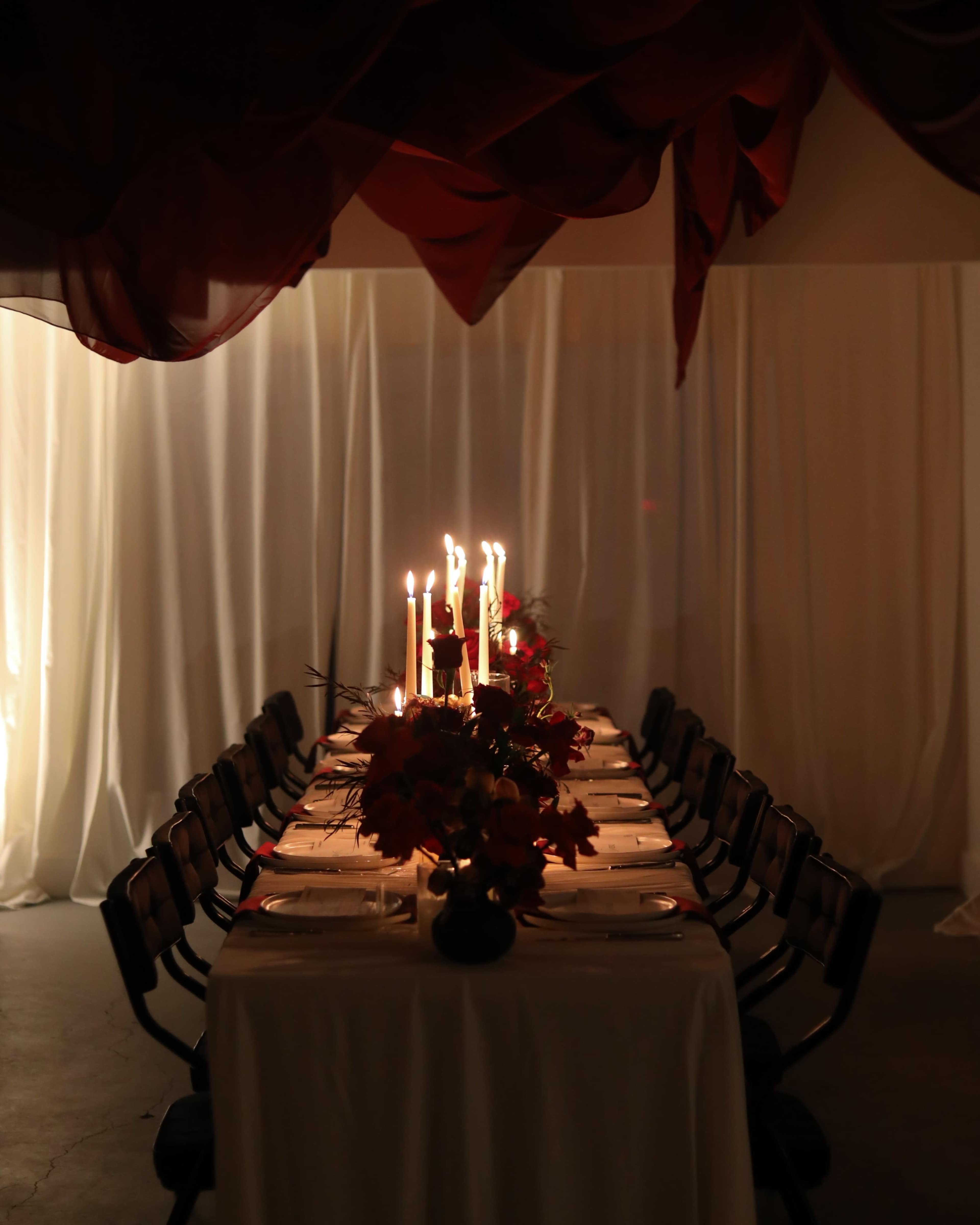 A long dining table is set with white tablecloths, candles, and floral arrangements, surrounded by chairs under draped fabric.
