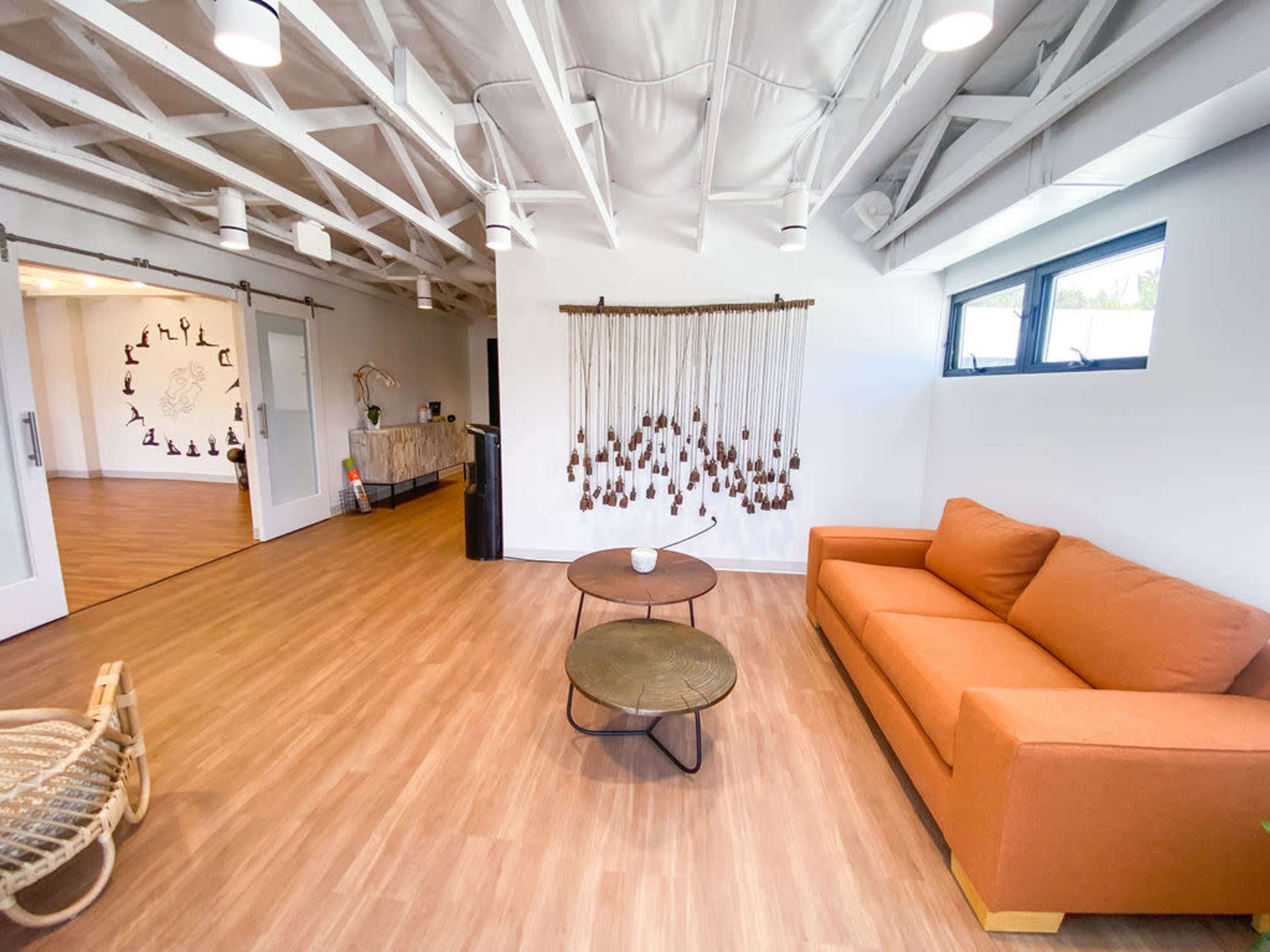 A modern interior space featuring an orange couch, two round tables, and a decorative wall hanging, with wooden flooring and exposed beams in the ceiling.