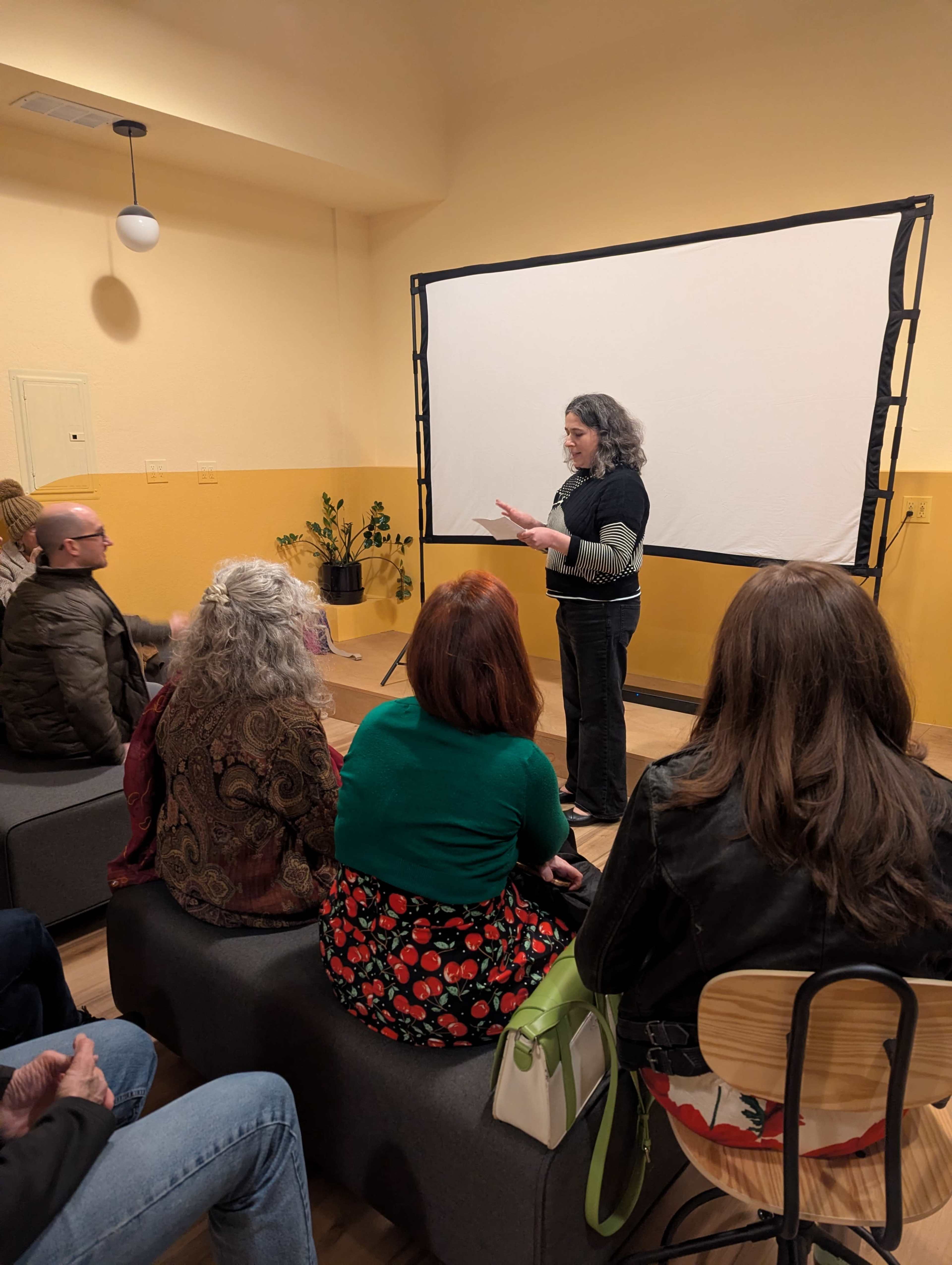 A speaker addresses an audience seated on modular furniture in a room with yellow walls and a projector screen.