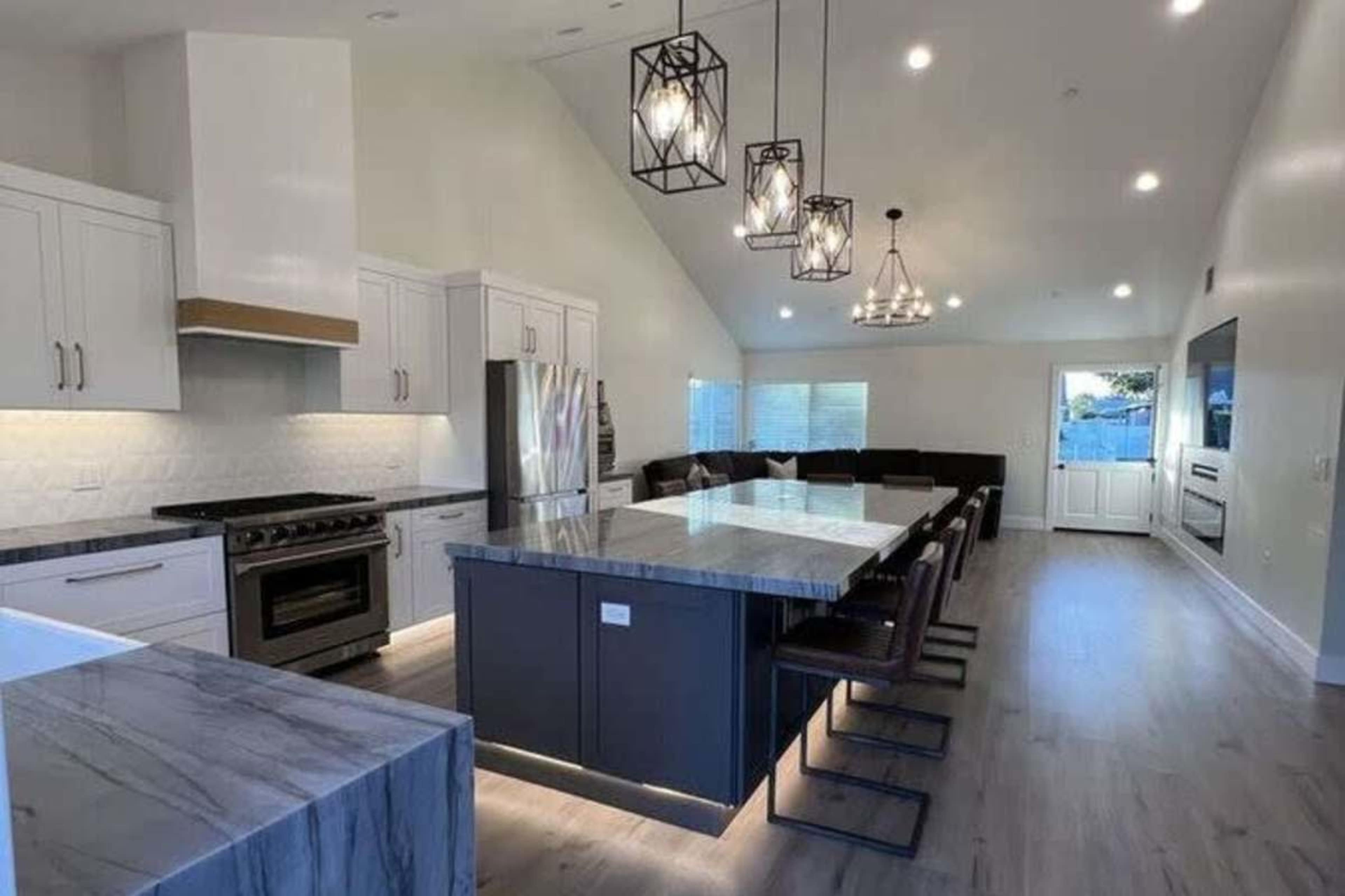 The image shows a modern kitchen with a large central island, stainless steel appliances, and a dining area with seating for several people.