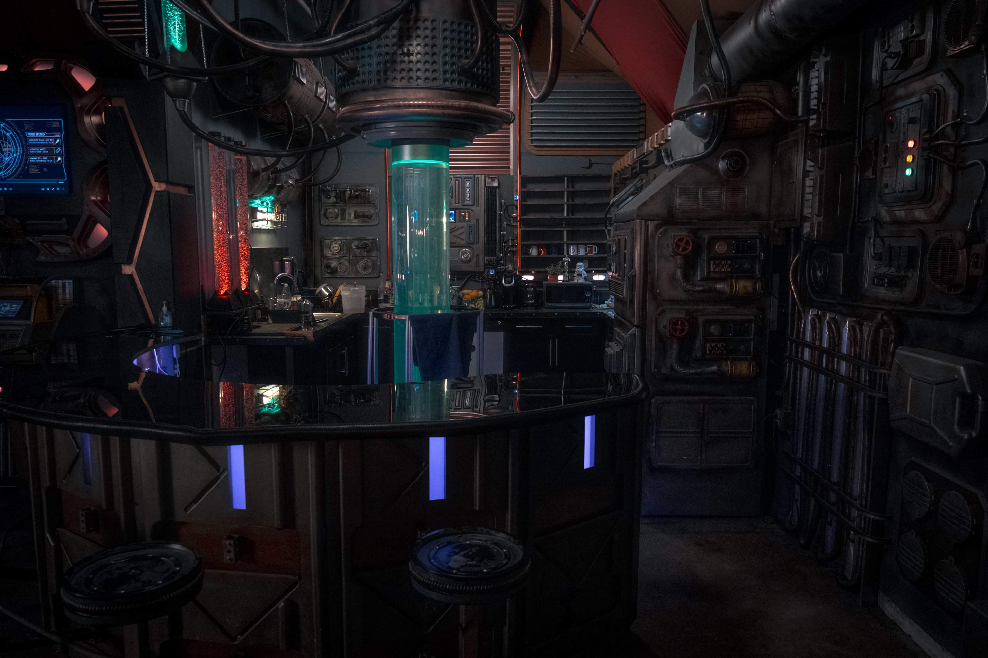 The image shows a dimly lit sci-fi bar with futuristic equipment, a glowing central column, and a high-tech counter surrounded by various gadgets and controls.