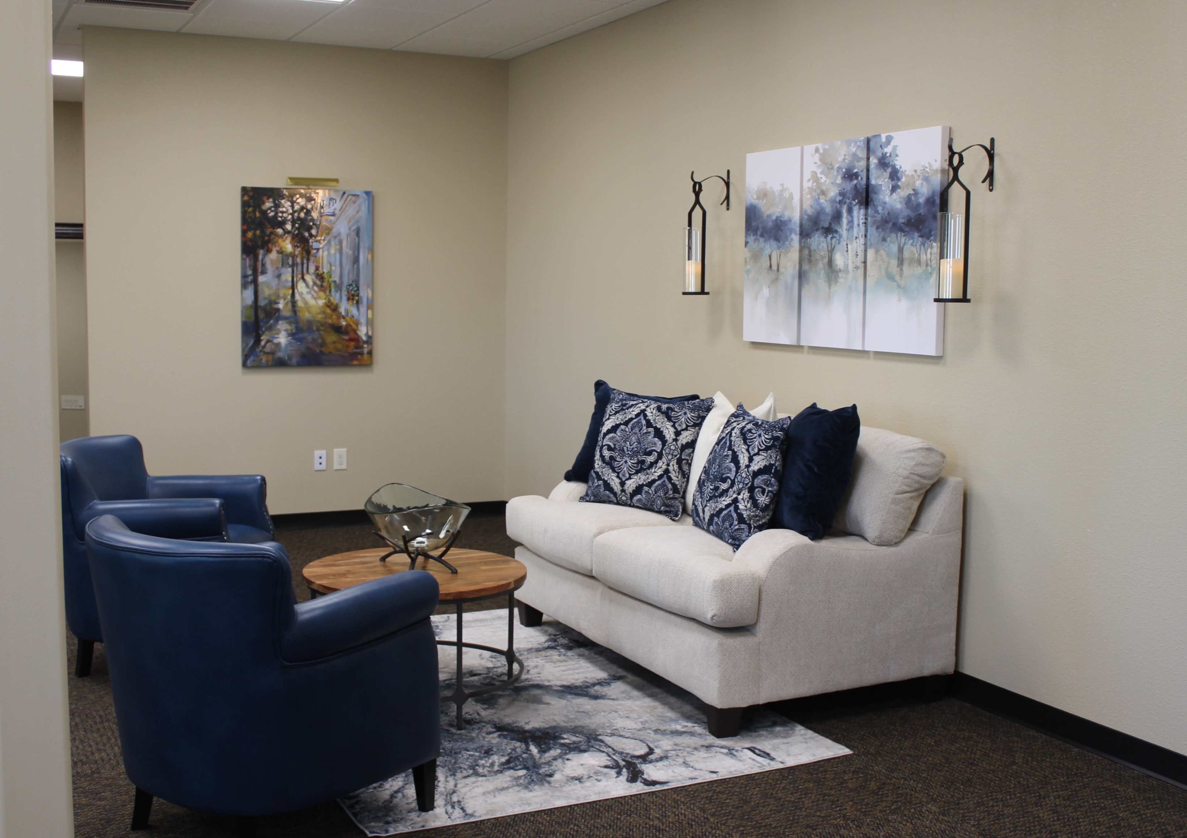 A cozy seating area featuring a light-colored sofa with decorative pillows, two blue armchairs, a round wooden coffee table, and artwork on the walls.